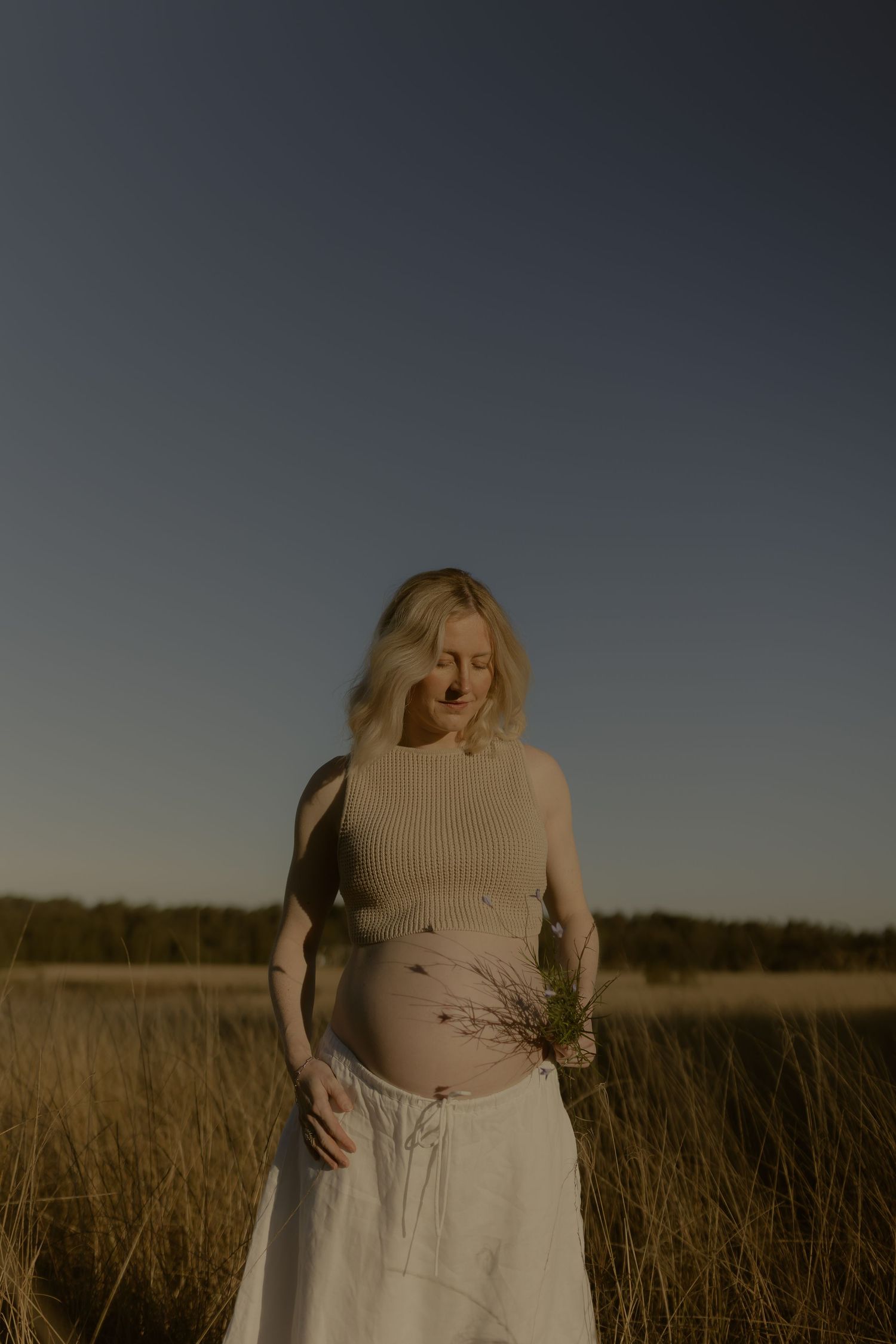 Pregnant woman in beige top and white skirt holding wildflowers in a golden field at dusk.