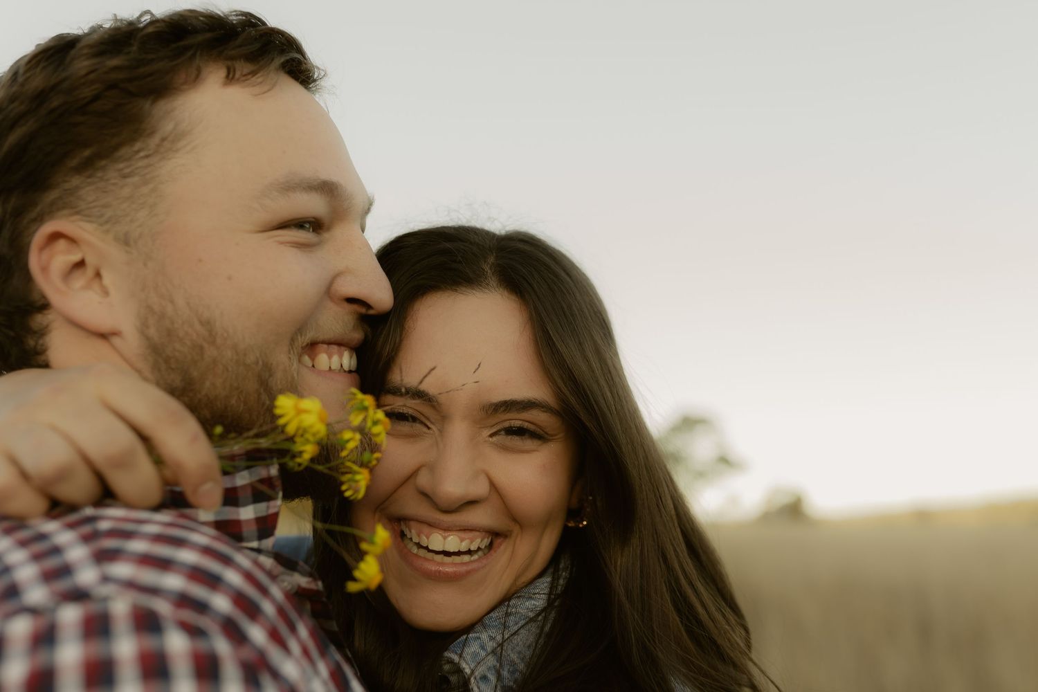 Happy couple laughing outdoors with man holding yellow wildflowers near womans face at sunset.