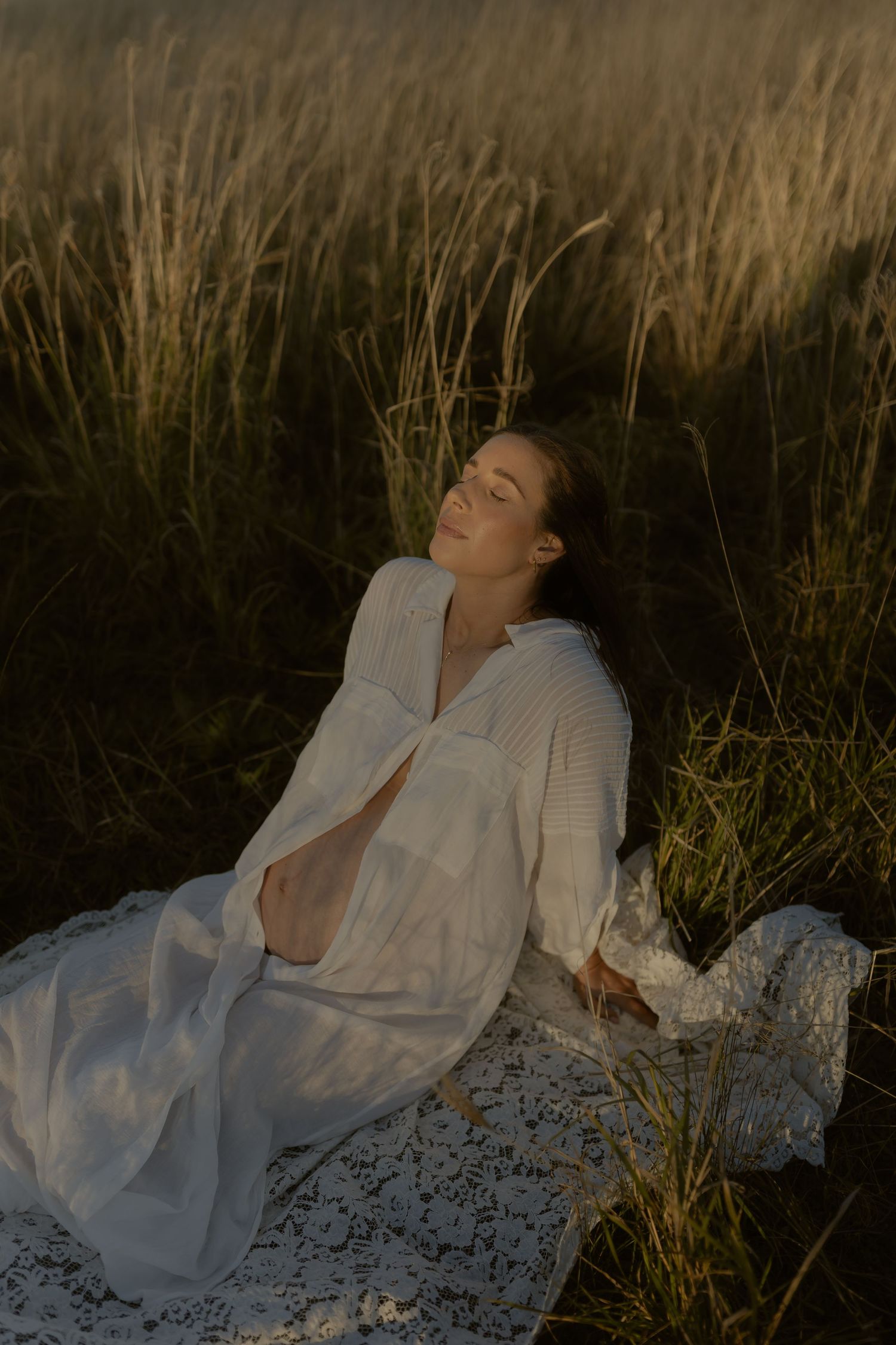Pregnant woman in white dress resting peacefully in golden grass field at sunset.
