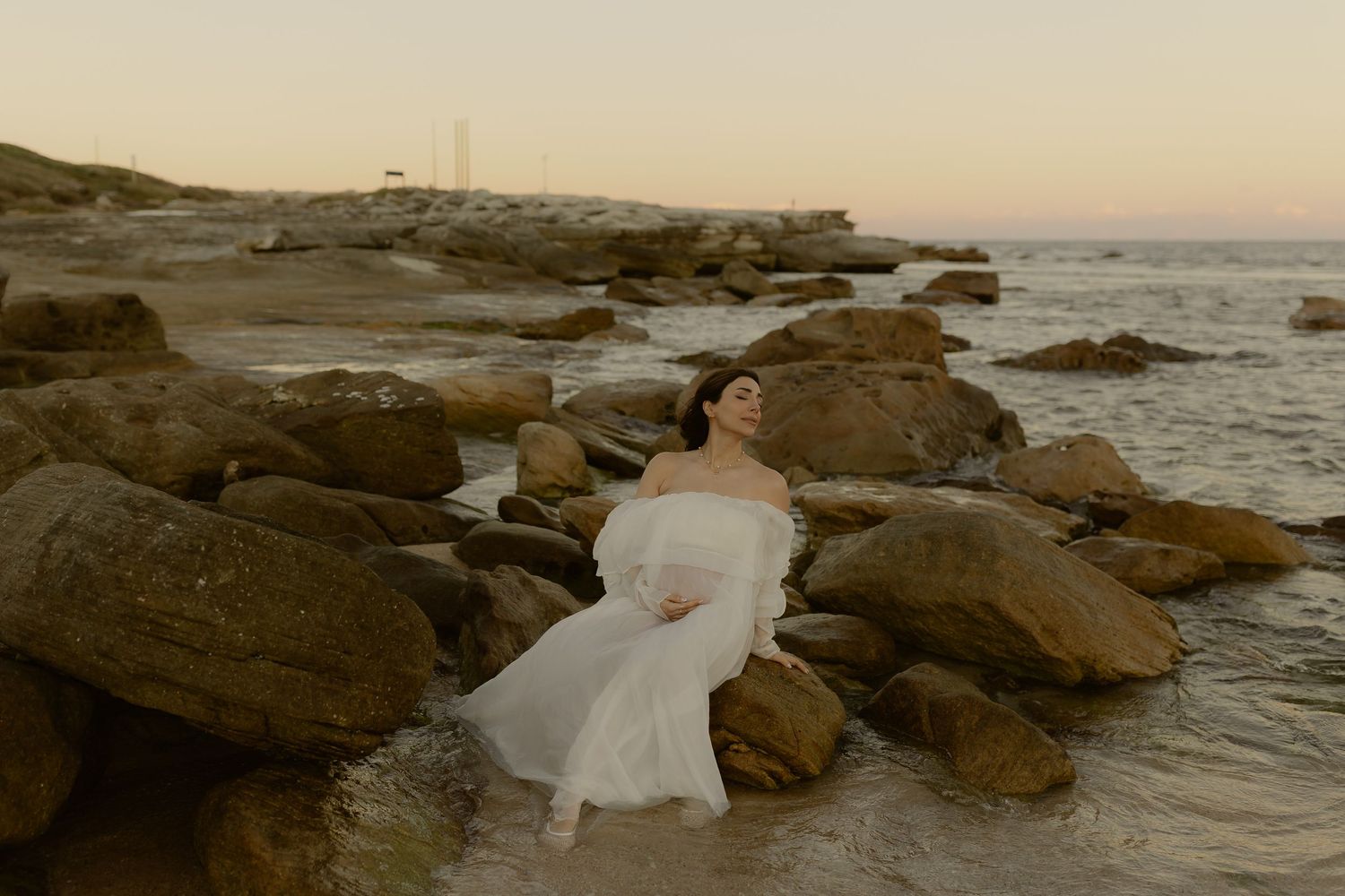 Bride in white off-shoulder gown reclining among ocean rocks at dusk.