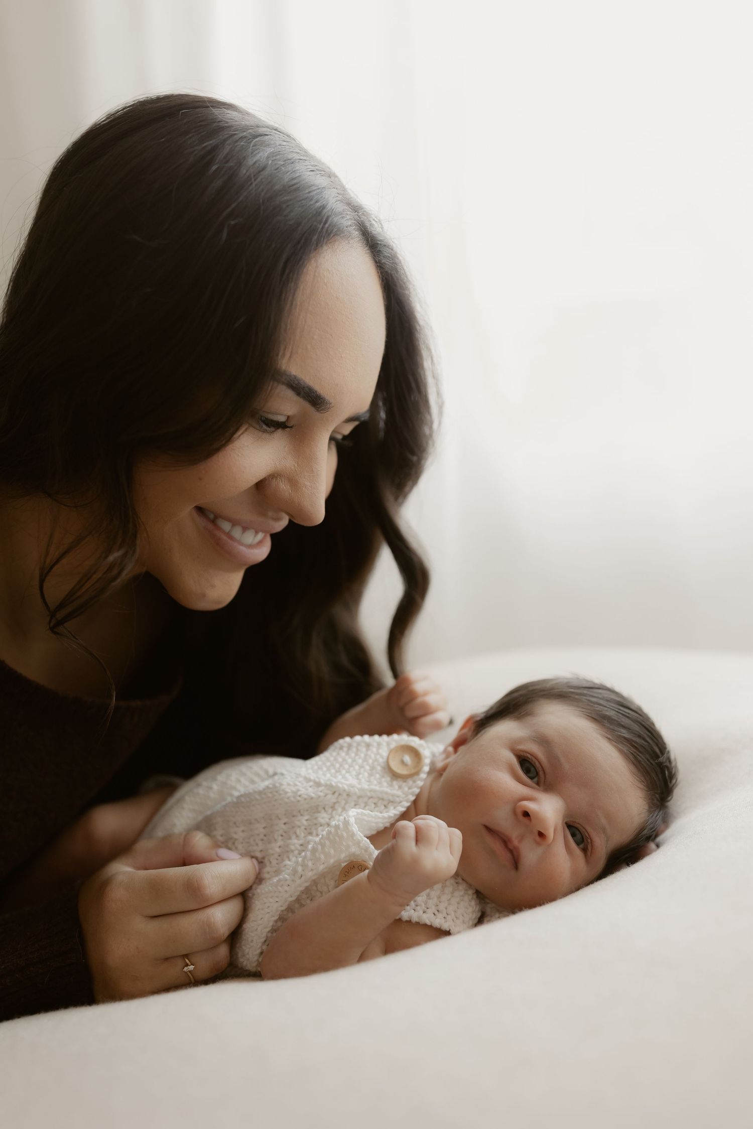 A smiling mother with dark hair gazes lovingly at her newborn baby dressed in a white knit outfit.