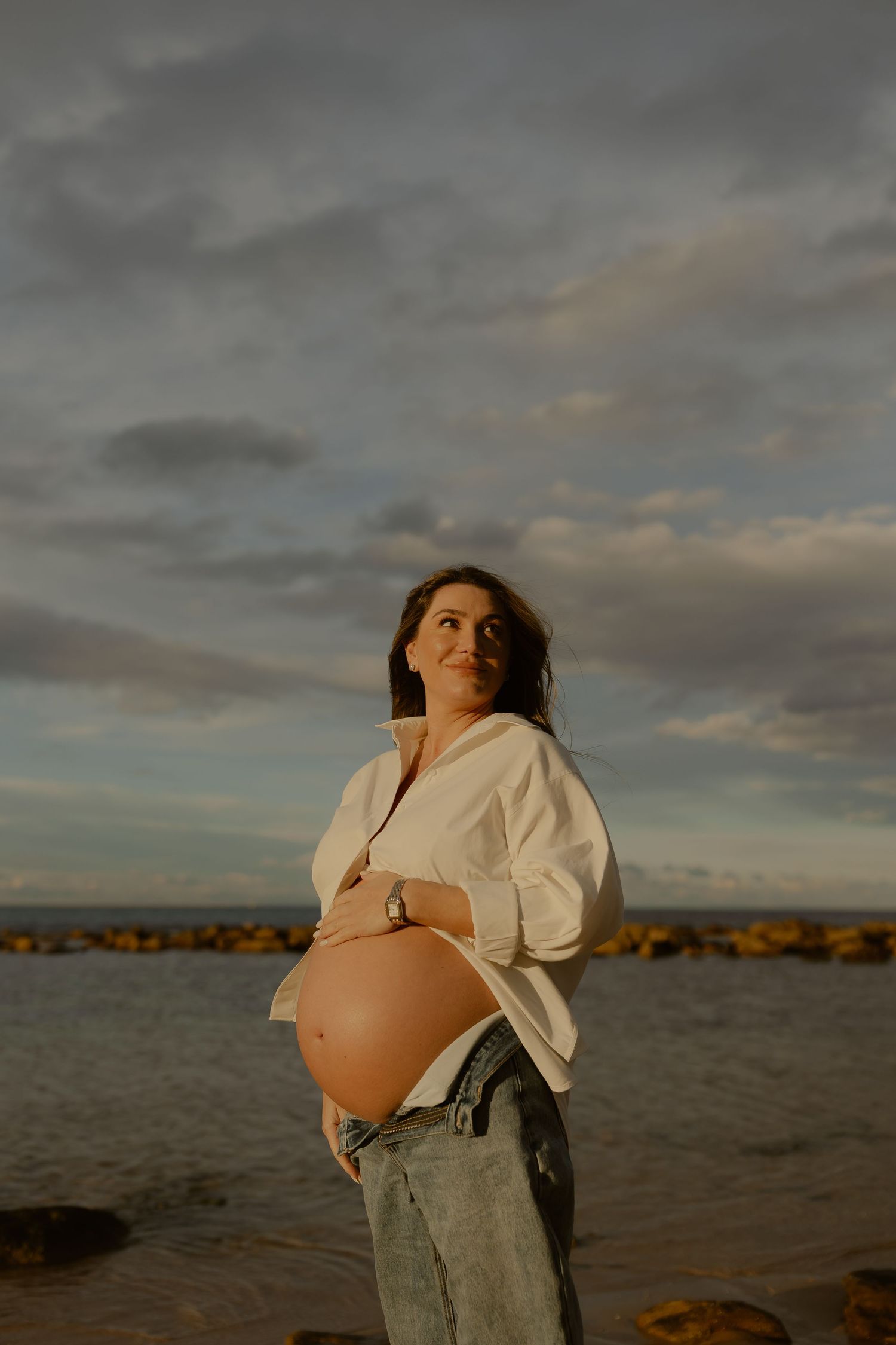 Pregnant woman in white shirt and jeans stands by rocky seaside under cloudy sky, cradling bare baby bump.