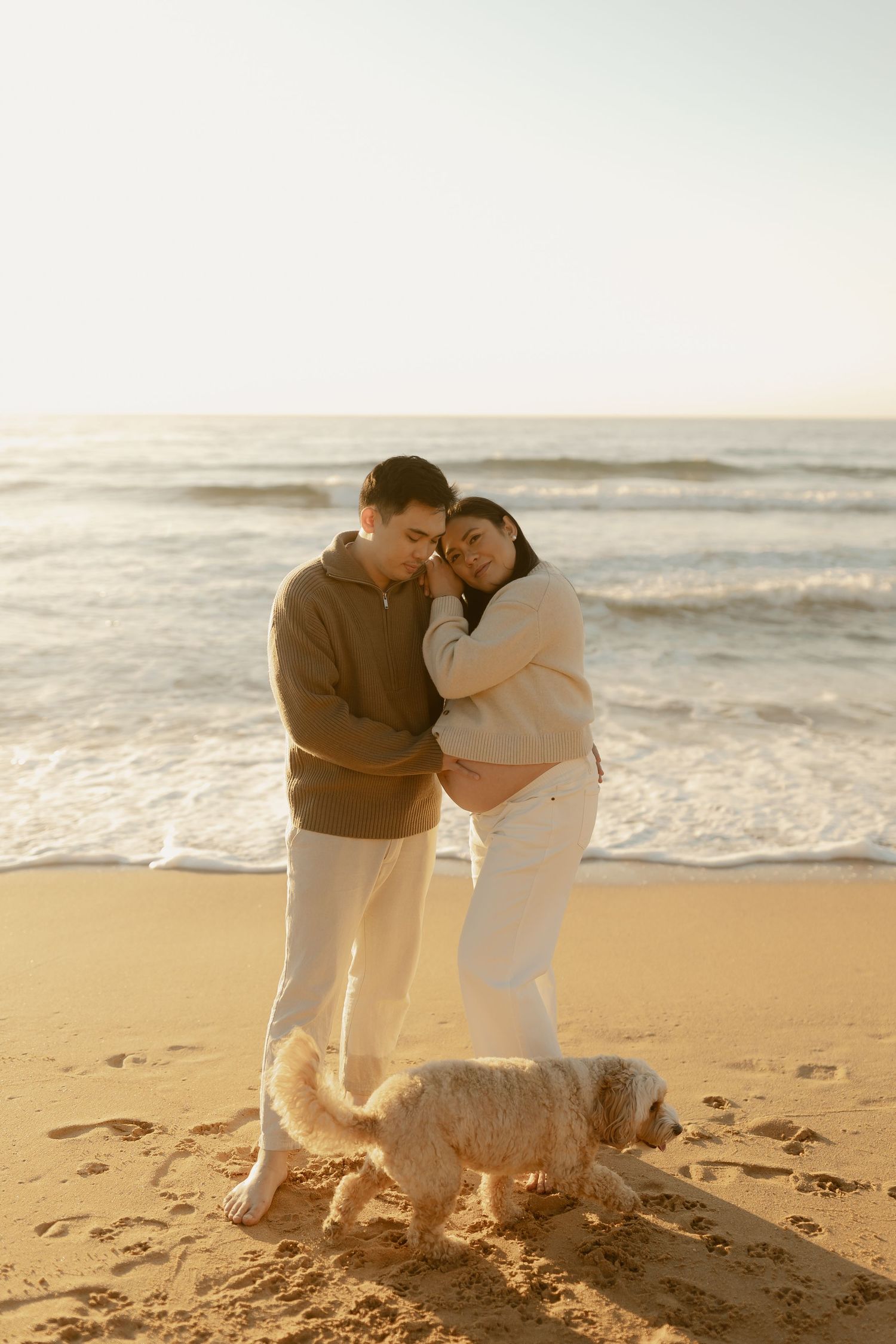 Couple embracing on a sandy beach at sunset with a fluffy white dog, ocean waves in the background.