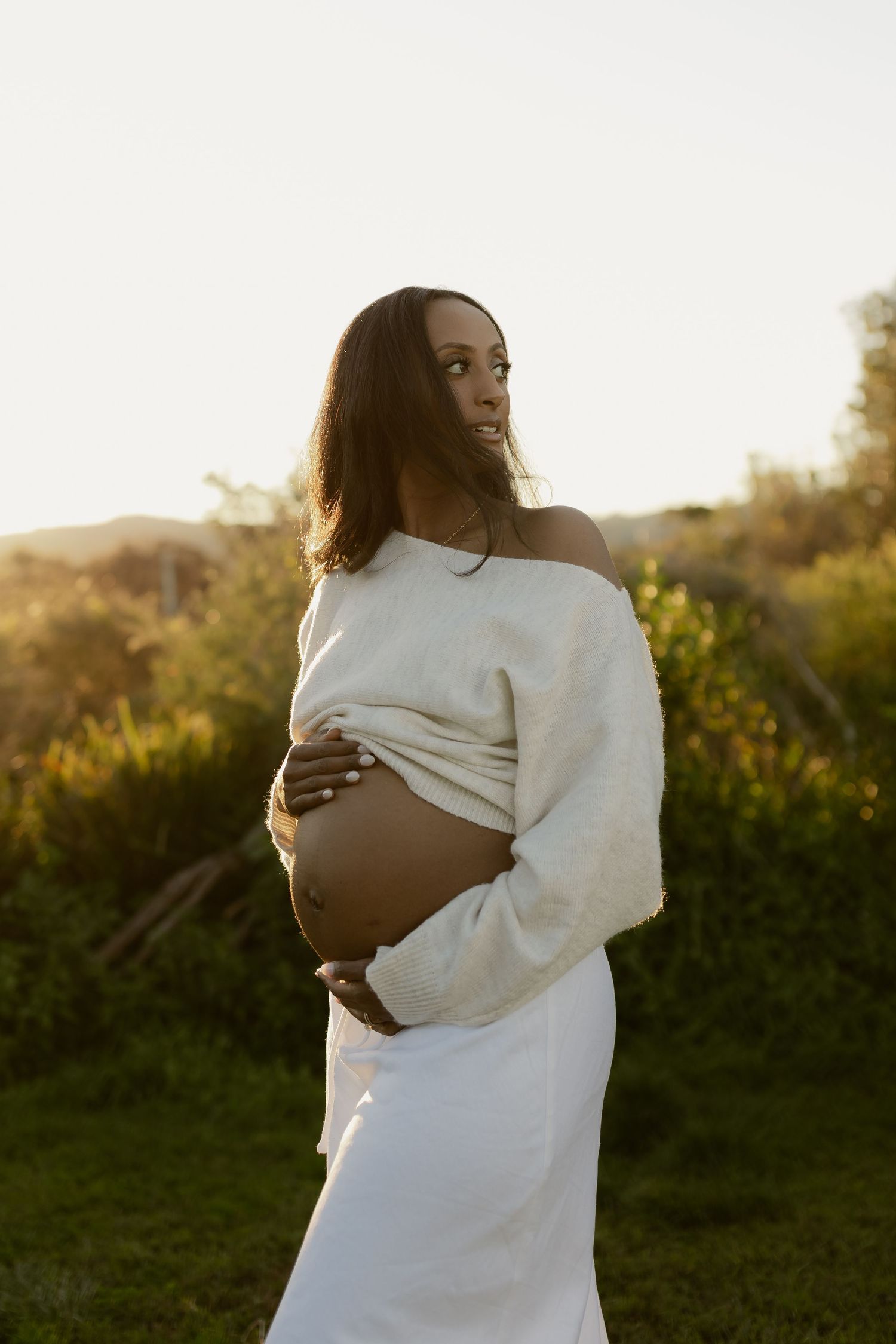 Pregnant woman in white skirt and sweater holding belly outdoors at golden hour sunset.