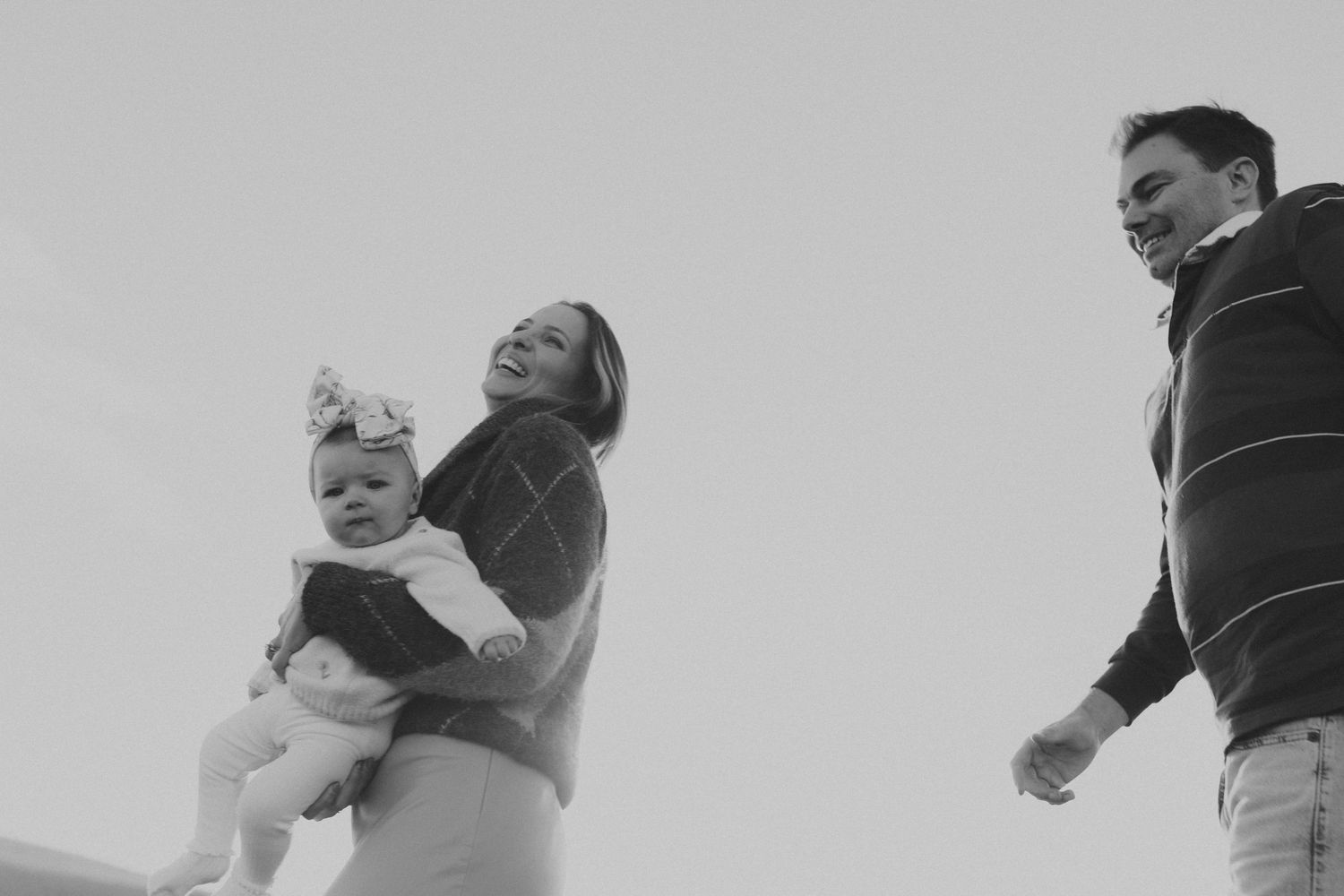 Black and white photo of happy parents lifting their baby girl outdoors against a bright sky.