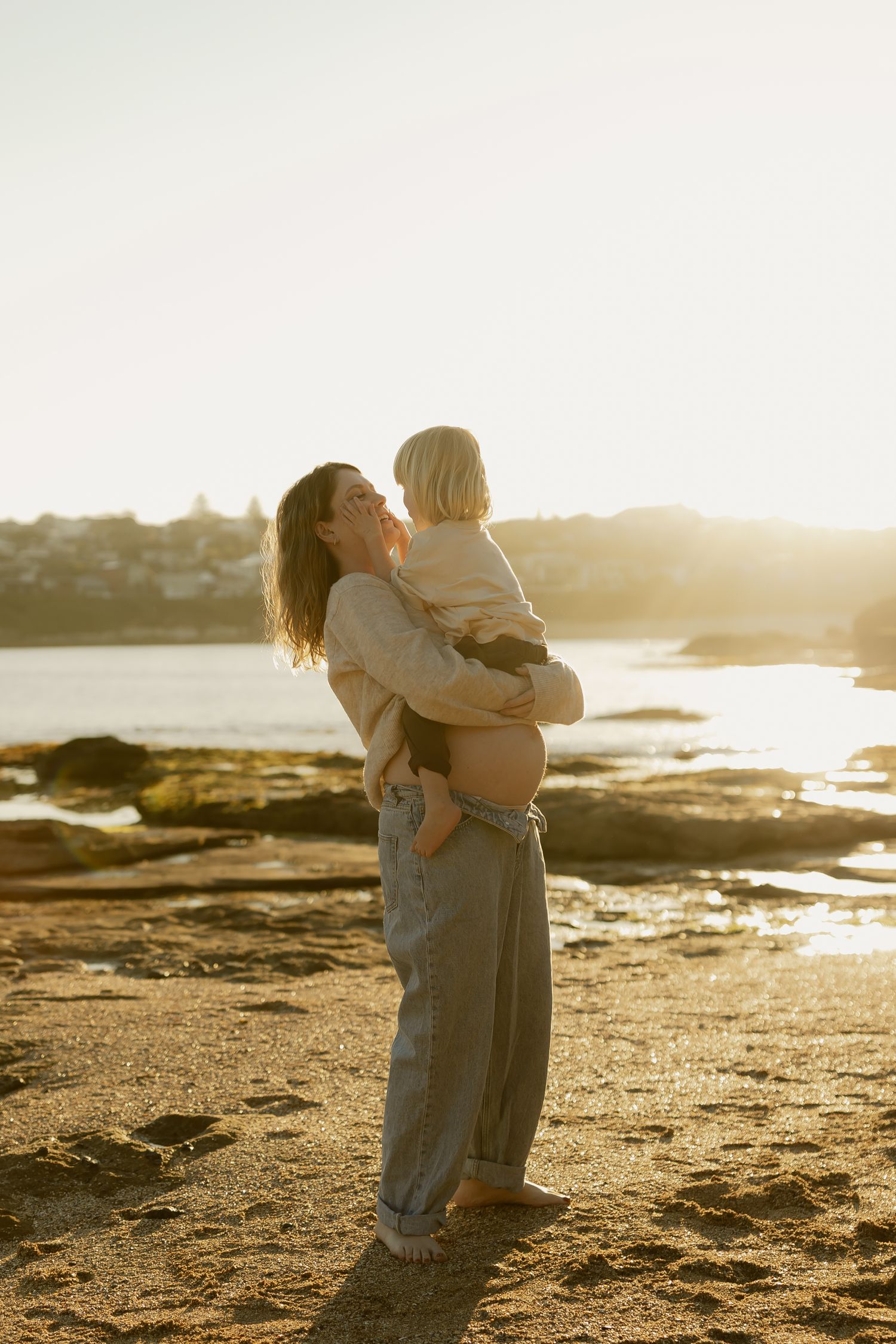 Mother lifting smiling toddler on sandy beach bathed in warm golden sunset light.