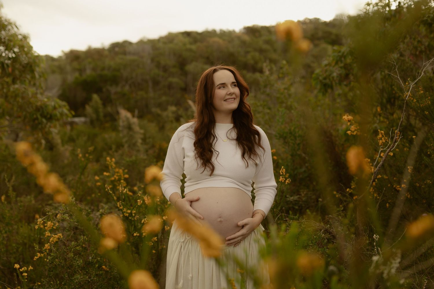 Smiling pregnant woman in white dress standing among yellow wildflowers and green bushes during golden hour.