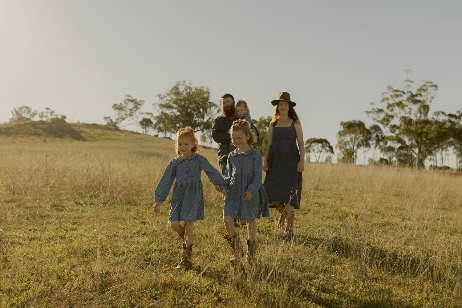 Family walking through golden grass field at sunset, two young girls in blue dresses holding hands with parents behind them.