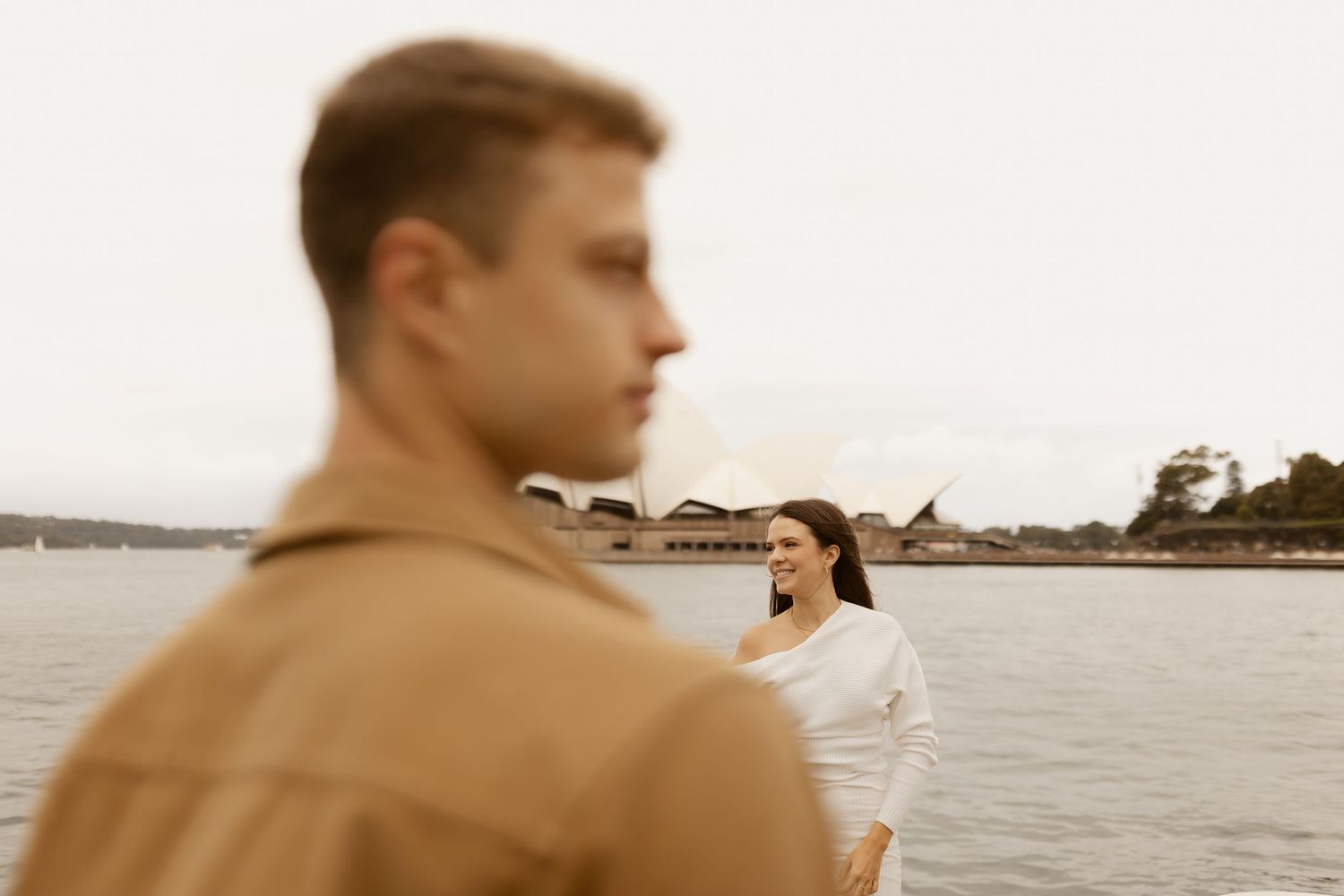 A couple stands by a waterfront on an overcast day, with a person in tan jacket in the foreground and water behind them.