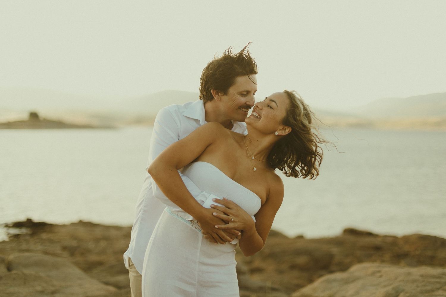 A romantic couple embraces and laughs together on a rocky beach during golden hour at sunset.