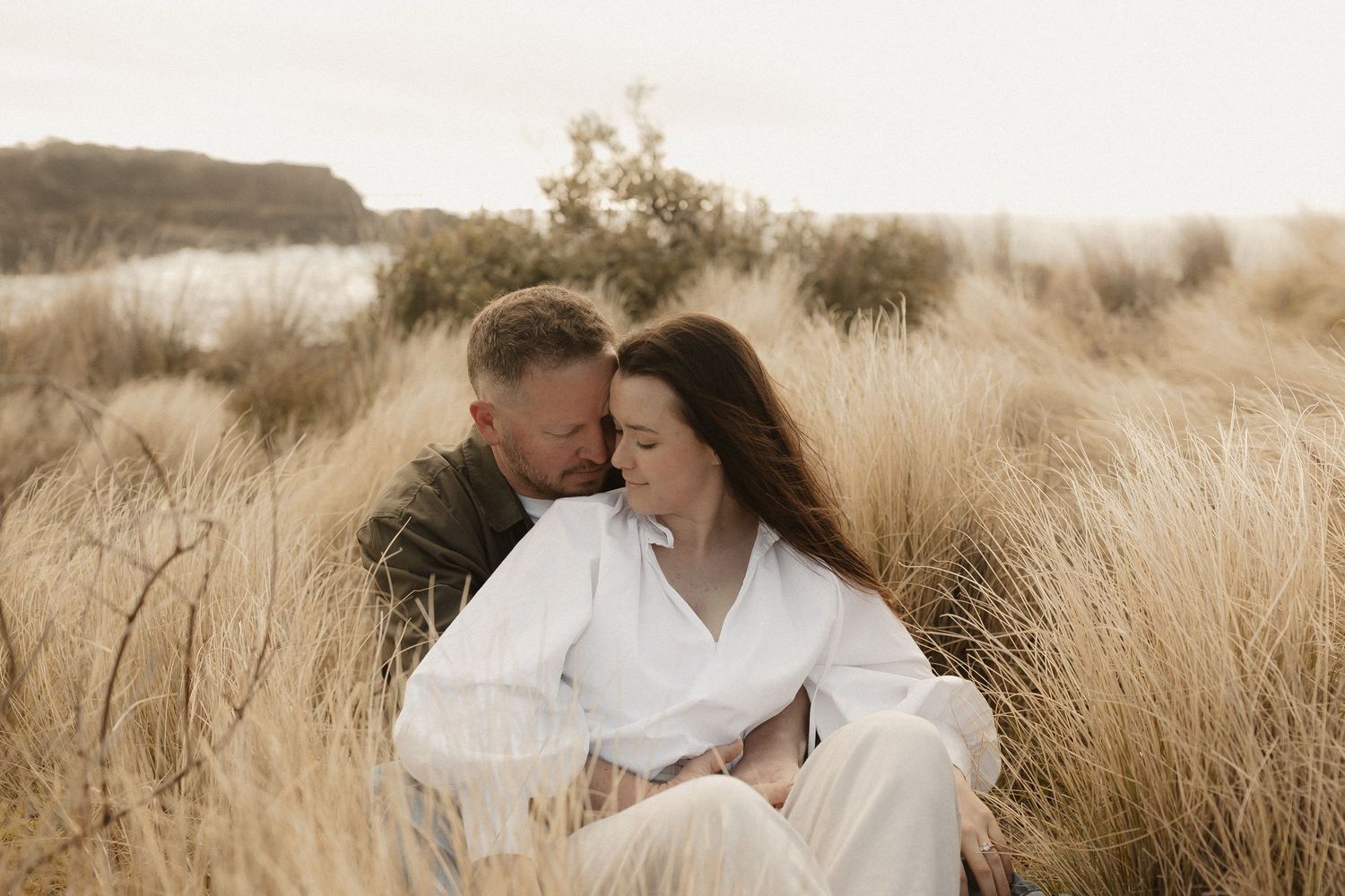 A couple embraces tenderly in a golden field of tall grass during sunset.