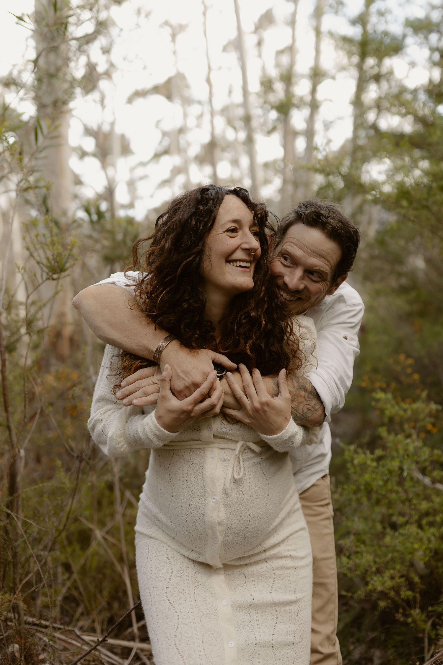 A joyful couple embraces in a forest setting, with the woman wearing a white maternity dress.