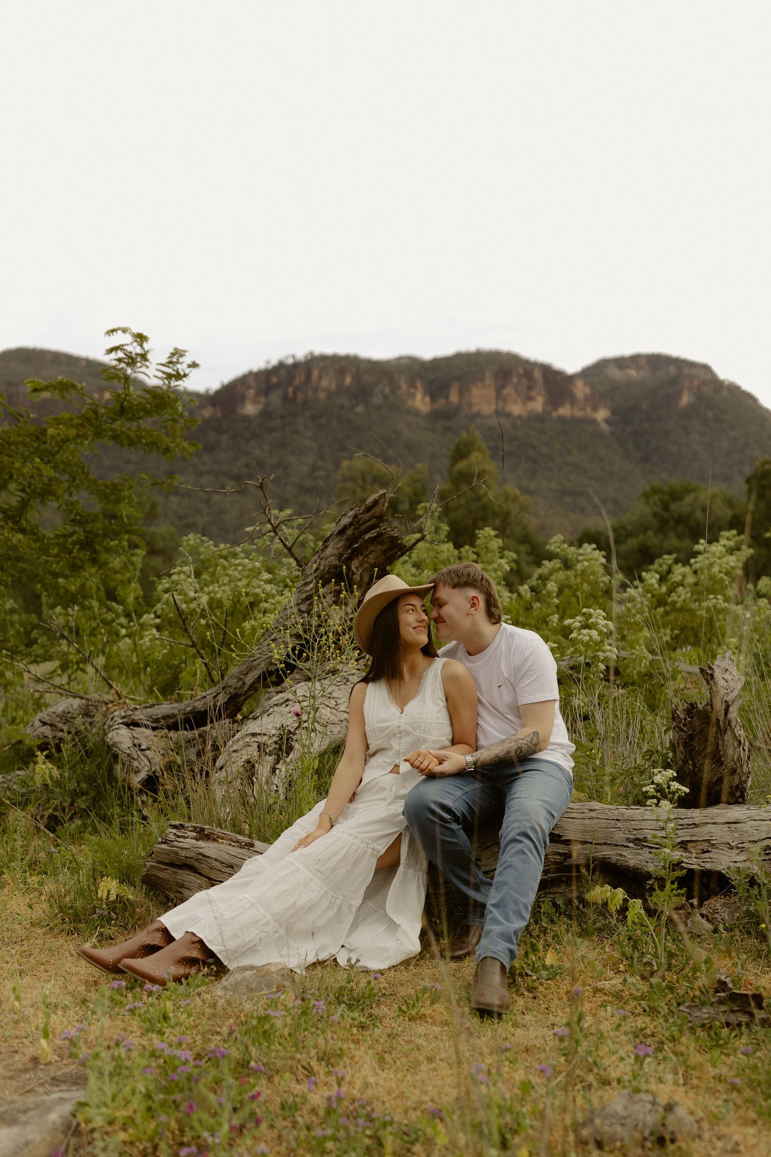 A romantic couple shares intimate moments on a grassy hillside with mountains in the background.