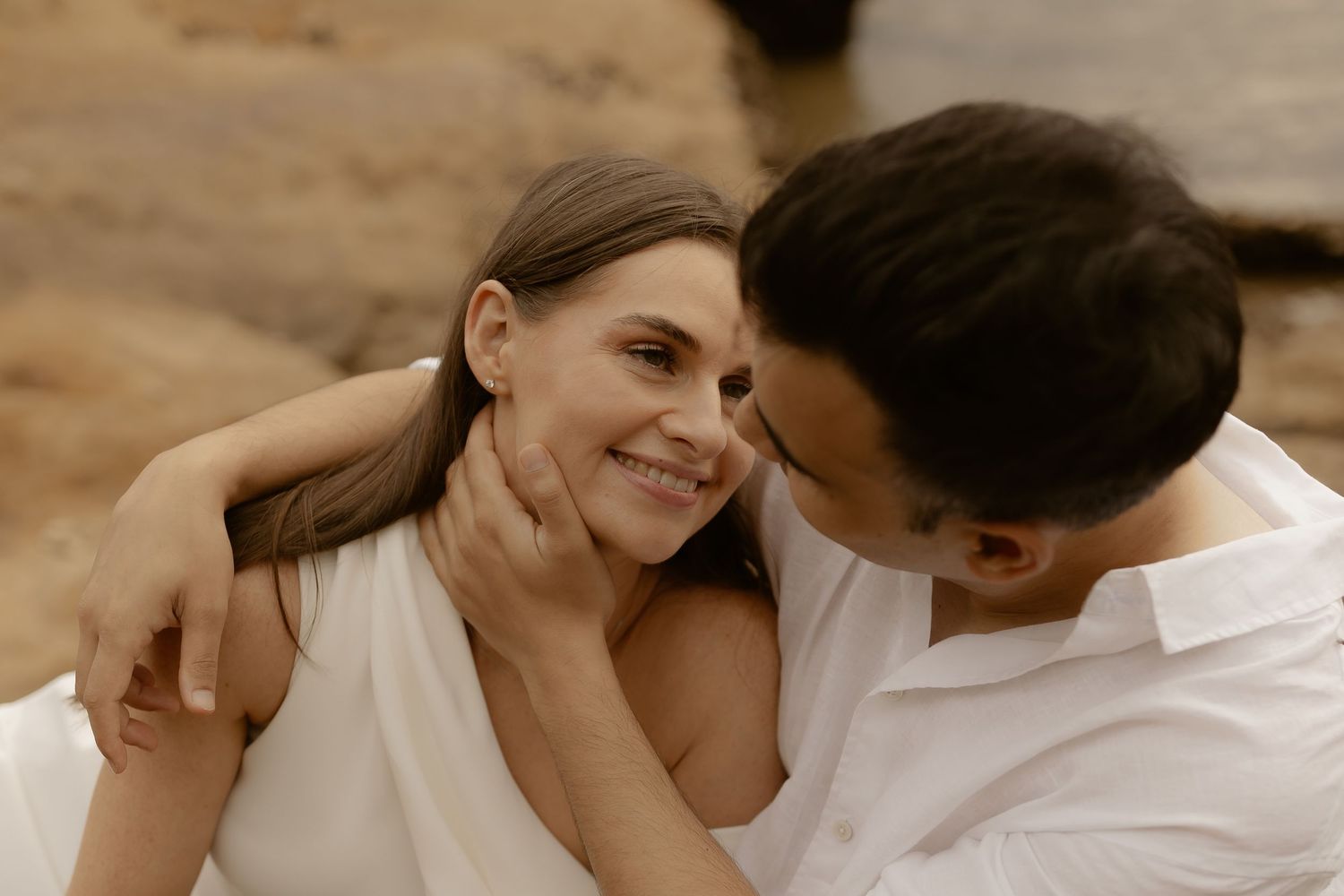 A romantic couple in white clothing embrace while sitting together during a warm outdoor photoshoot.