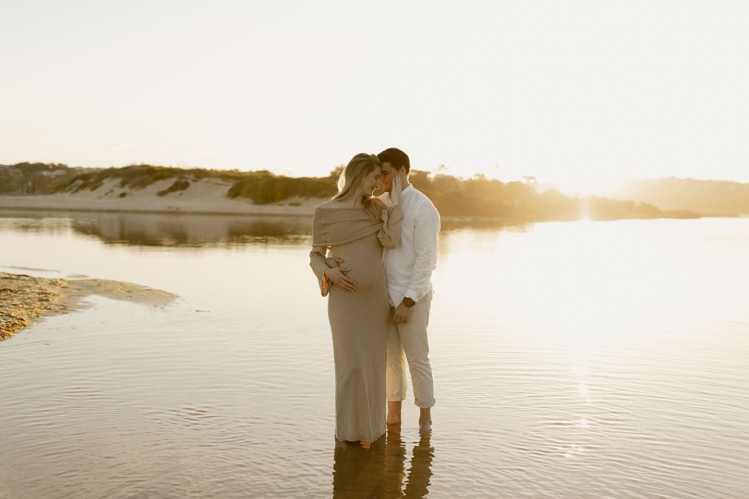 A couple embraces on a beach at sunset with golden light reflecting off the water.