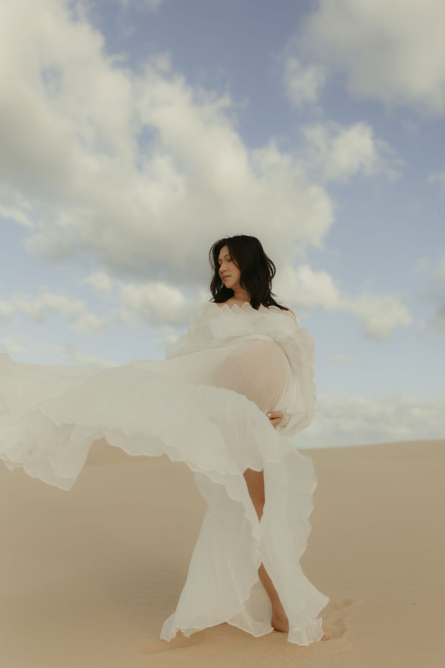 A figure in a flowing white dress dances gracefully across desert sand dunes under a cloudy sky.