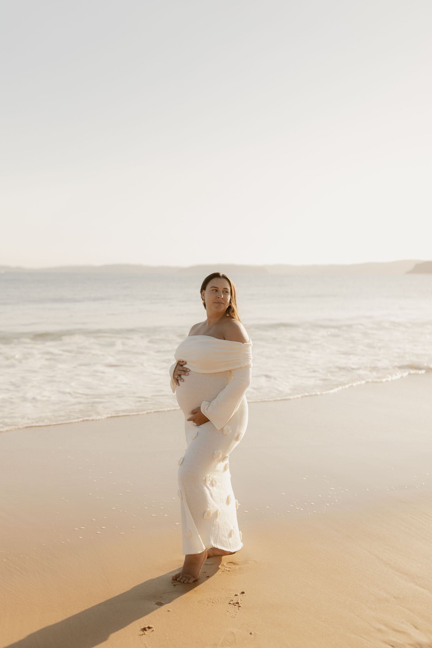 Figure wrapped in white fabric poses dramatically on a sunlit sandy beach.