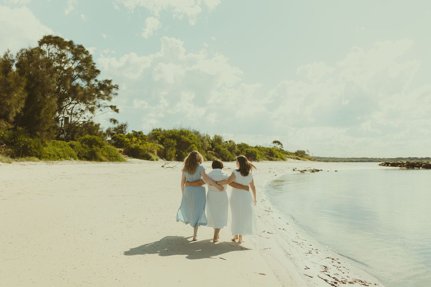 People in white clothing walk along a pristine sandy beach with turquoise waters and blue sky in the background.