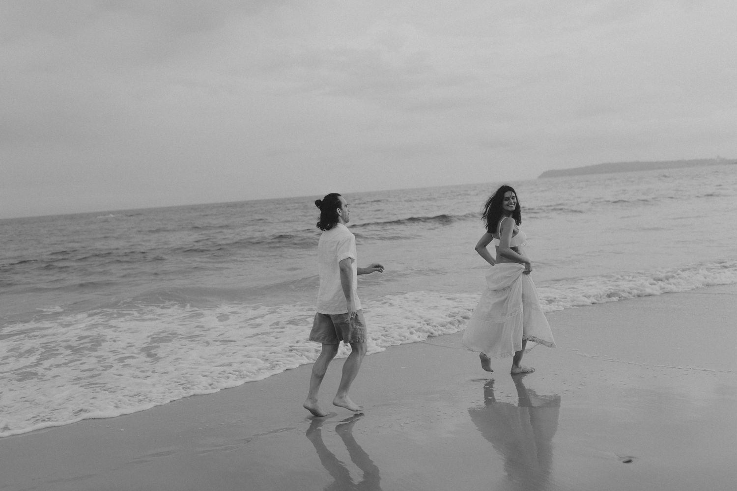 Two people playfully run along the shoreline in black and white.
