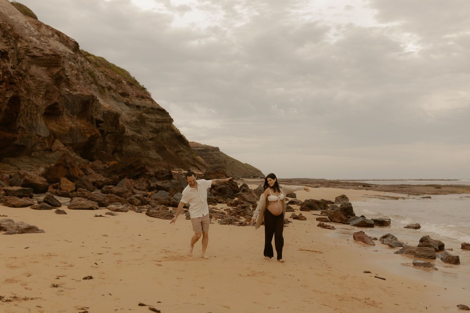 People walking along a dramatic coastal beach with large rock formations and overcast sky.