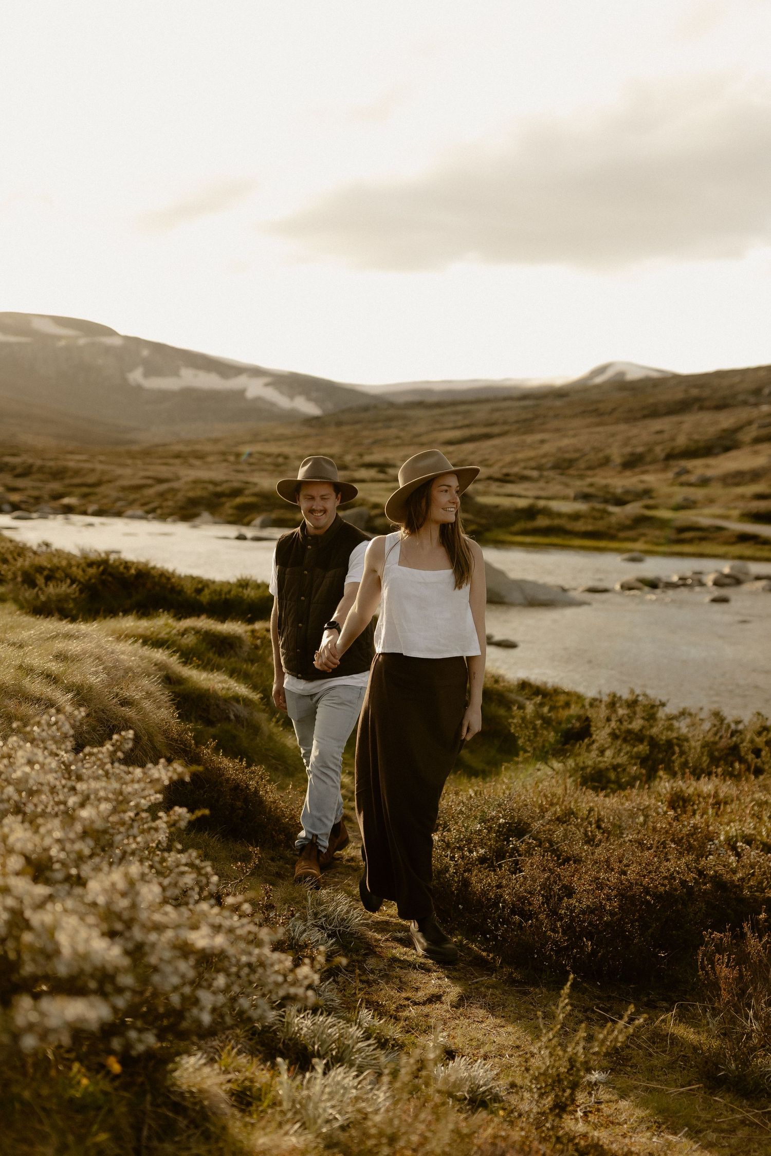 A couple walks together through a scenic mountainous landscape with a lake in the background at sunset.