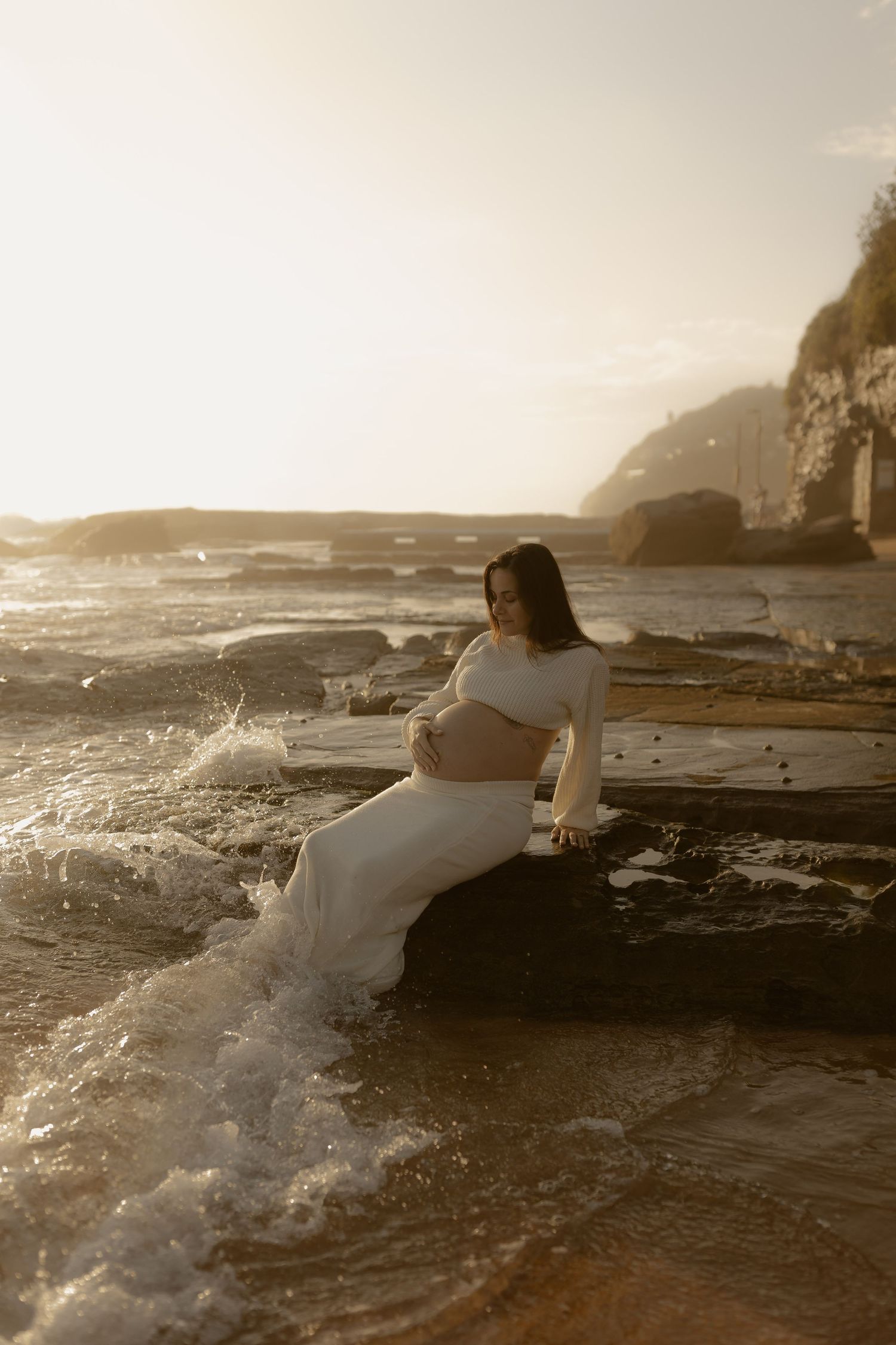 Waves crash against rocky coastal ledges at sunset as someone in white clothing sits near the ocean's edge.