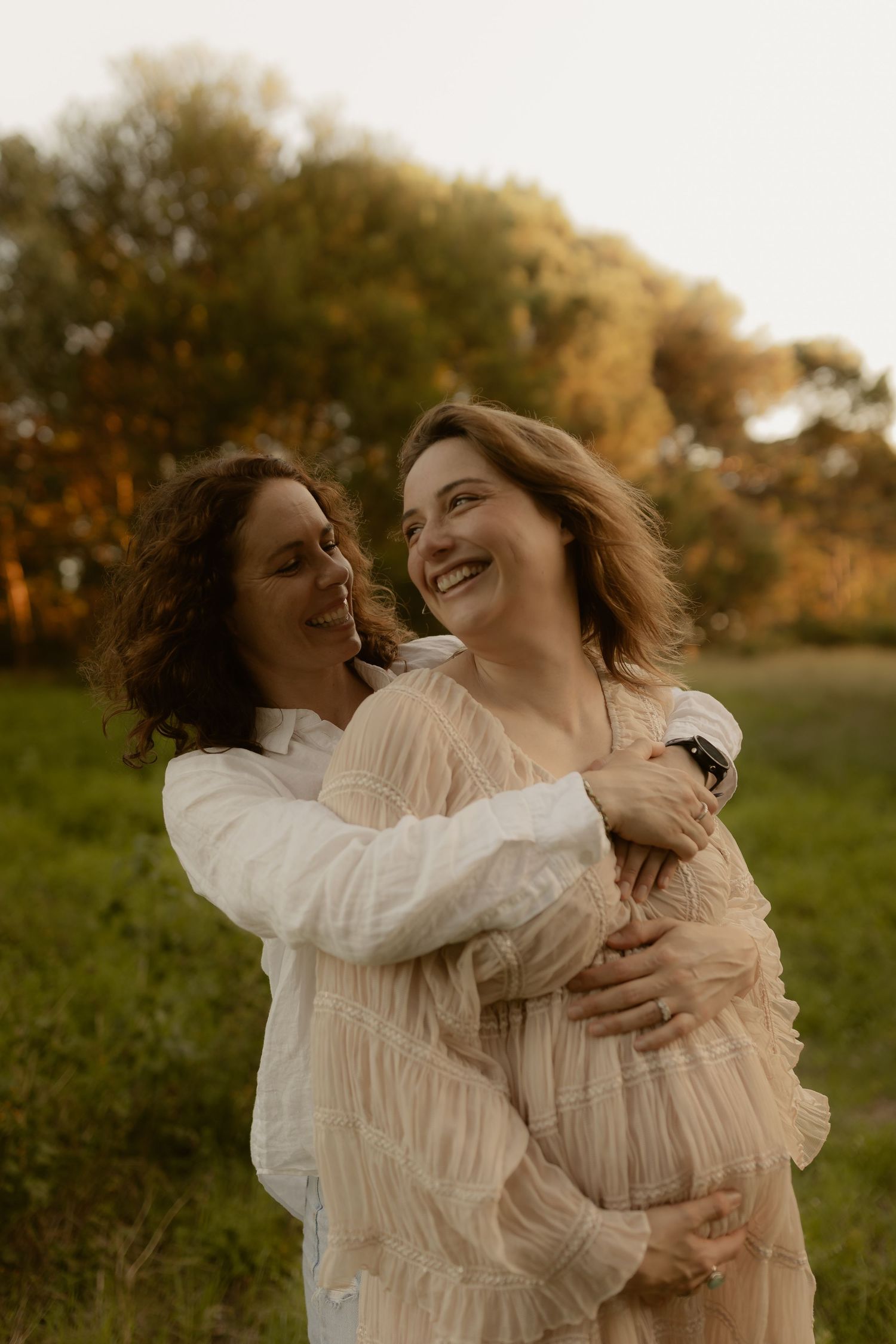 Two people in white dresses sharing joyful embraces in warm sunset light.