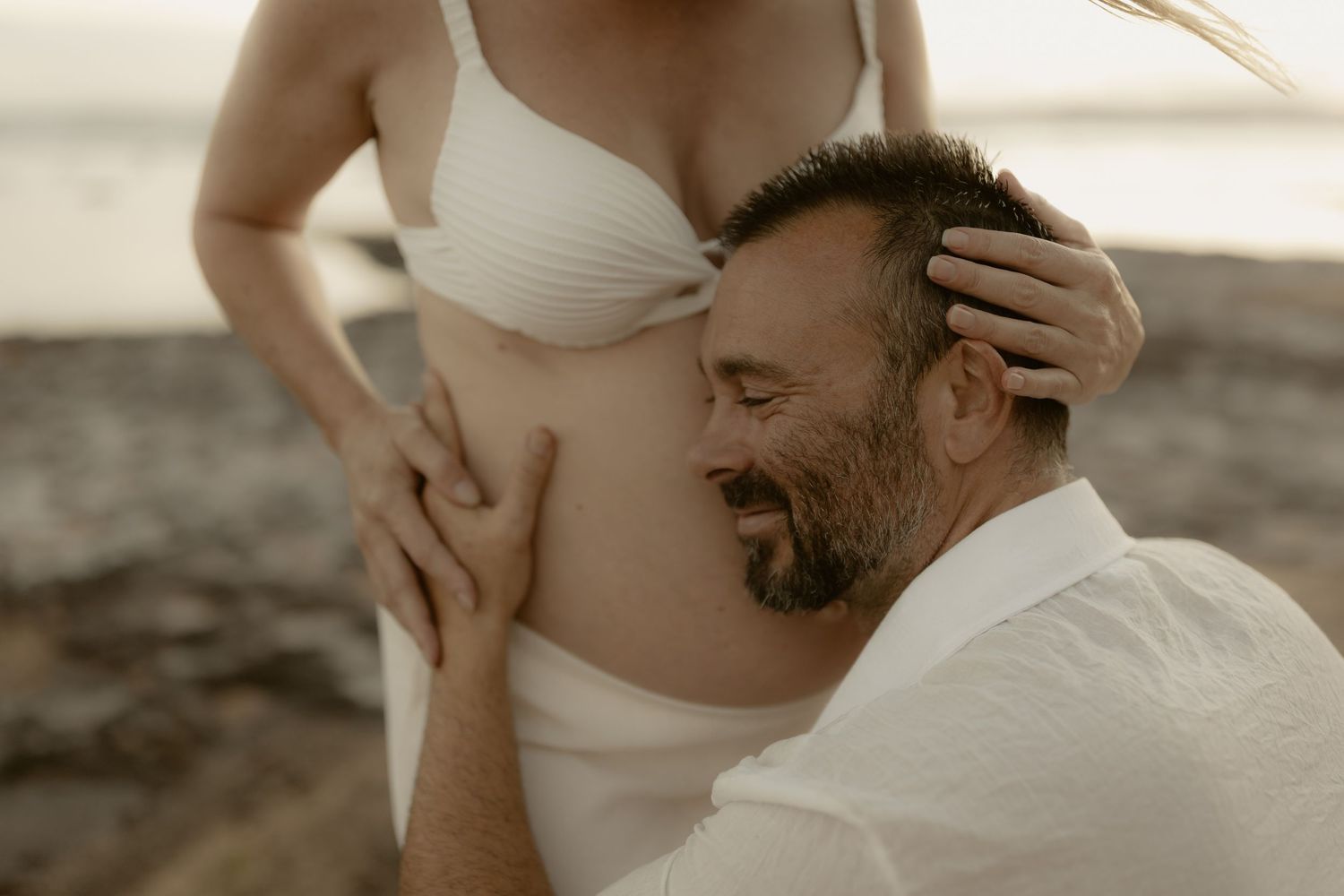 Intimate beach photoshoot with couple in white attire during golden hour against rocky coastal backdrop.