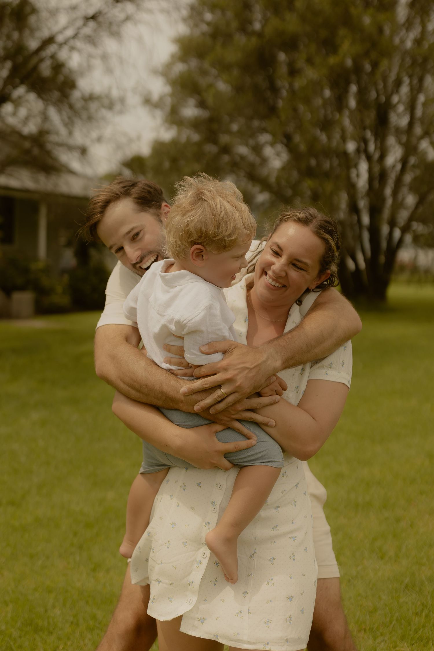 A tender family moment captured in a sunlit grassy field with trees.