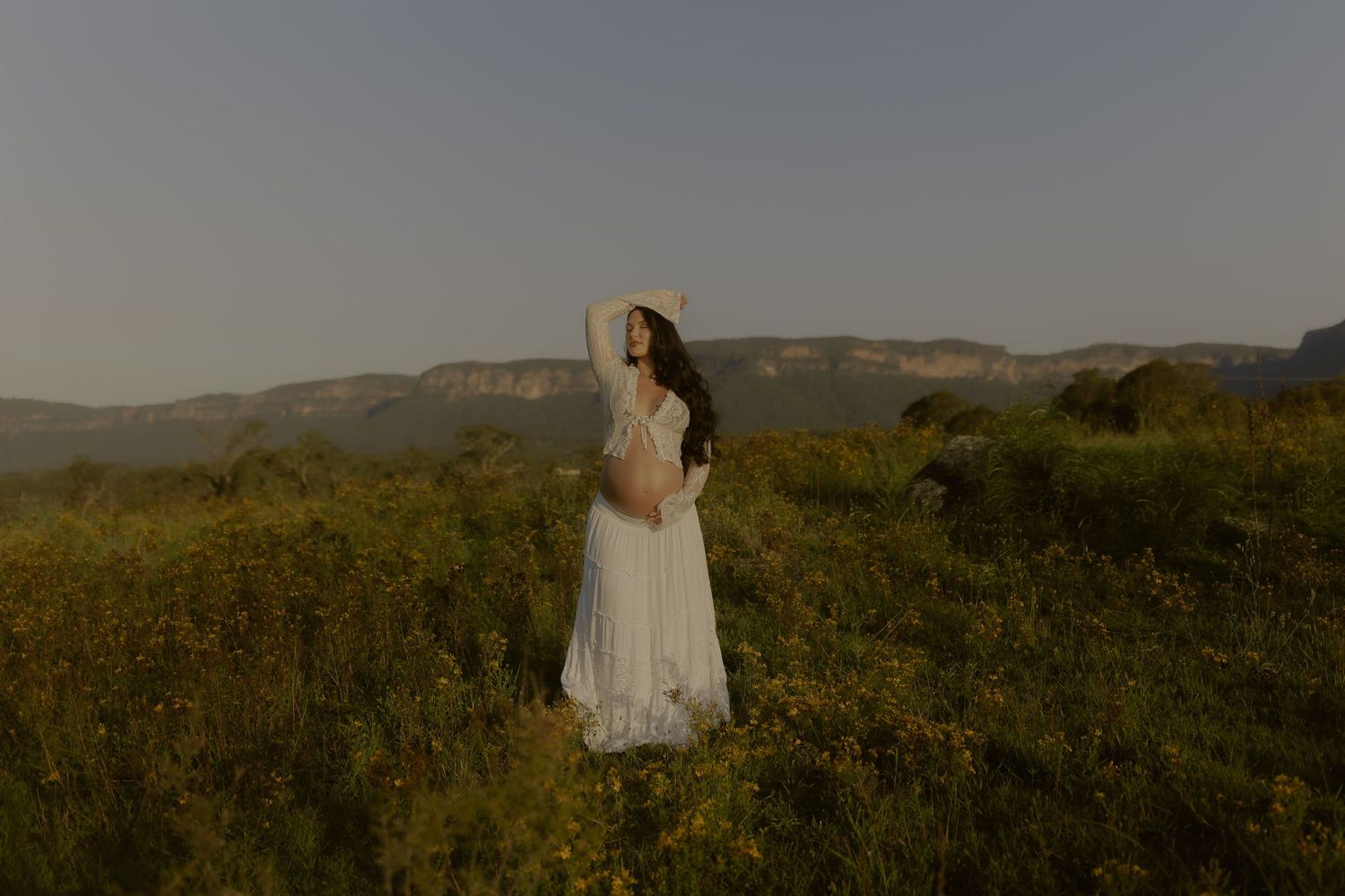 Figure in white dress walks through tall grass on a hilltop during sunset.