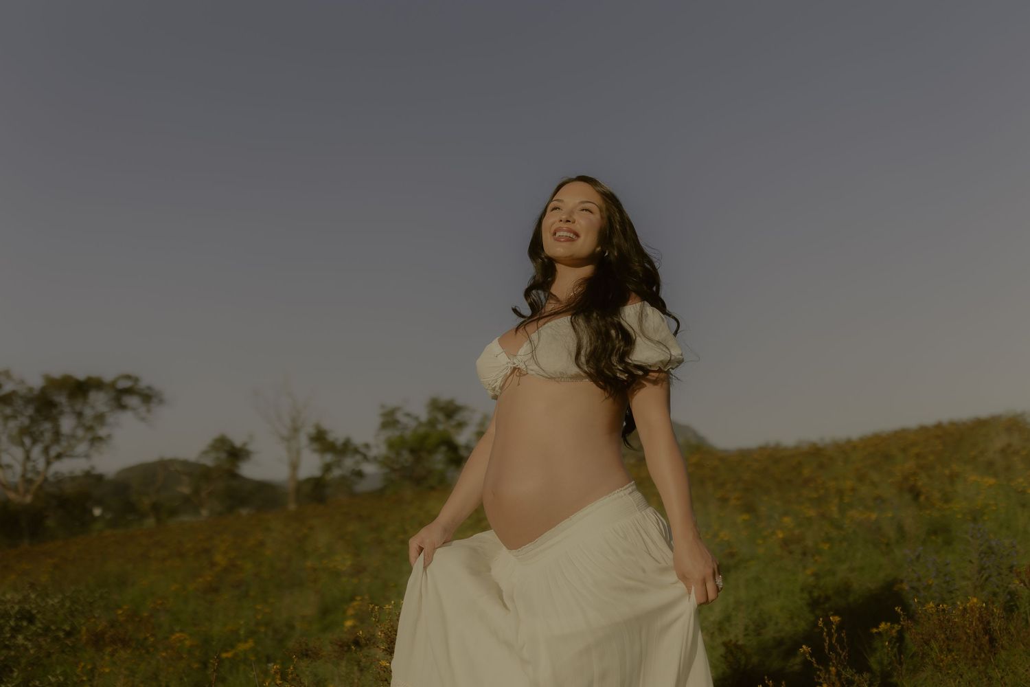 Expectant mother in white dress poses gracefully in golden sunlit meadow.