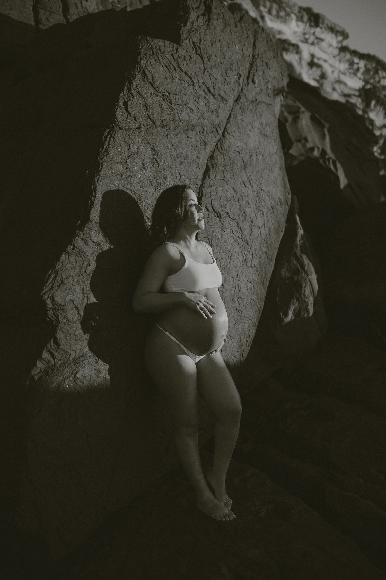 Black and white artistic maternity portrait featuring an expectant mother posing against a dramatic rocky cave wall.