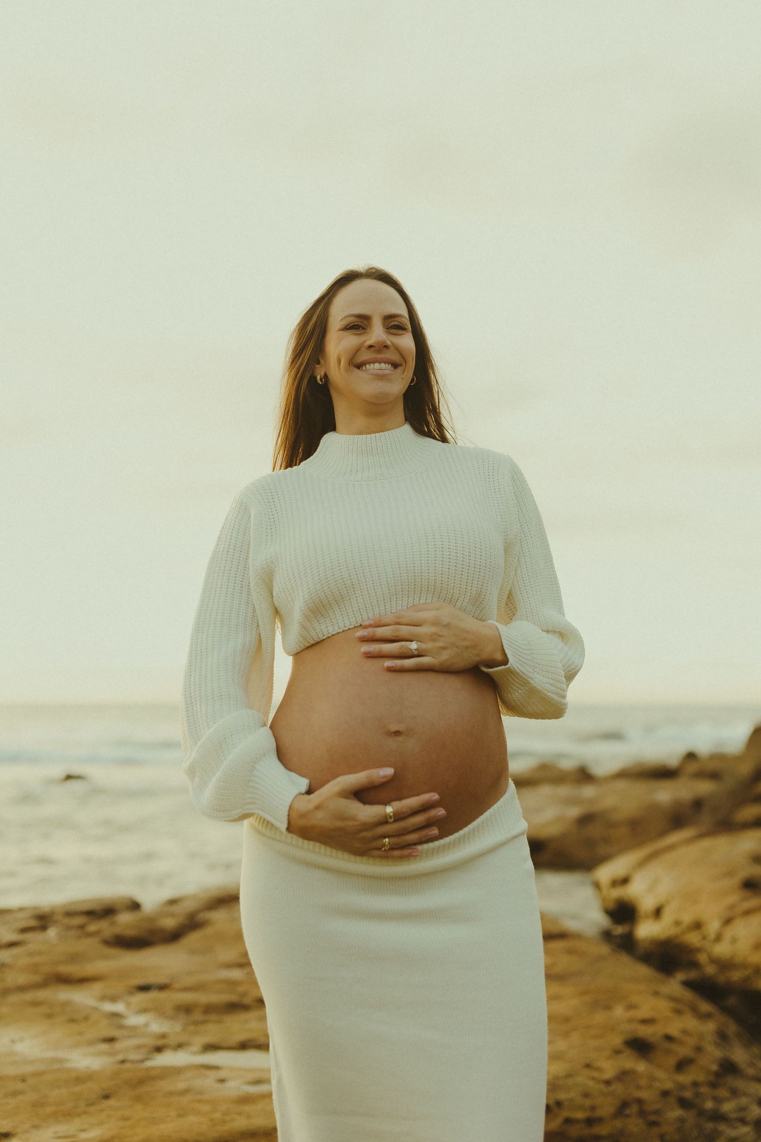 A maternity photoshoot on a rocky beach with someone wearing a white knit dress.