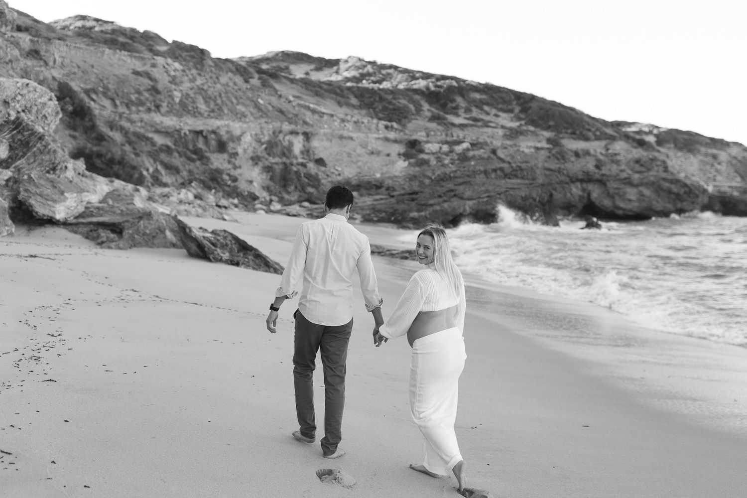 A couple strolls together on a secluded beach with dramatic cliffs in the background in black and white.