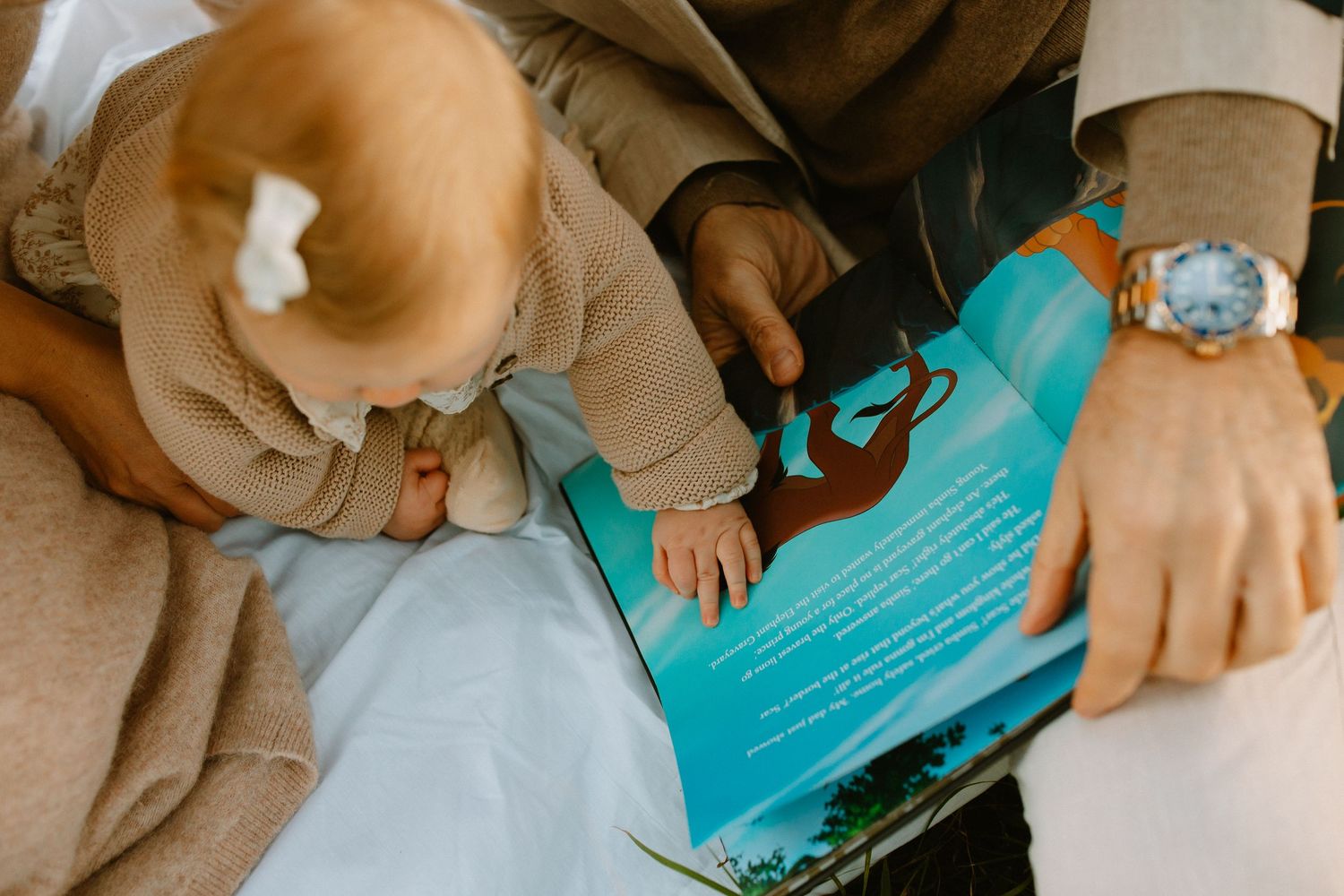 Close-up view of hands holding a turquoise book while a baby in beige clothing reaches towards it.