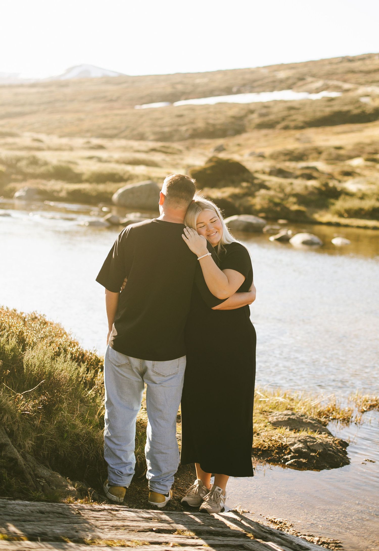 Couple stands together in silhouette against sunlit lake and rolling hills during golden hour.