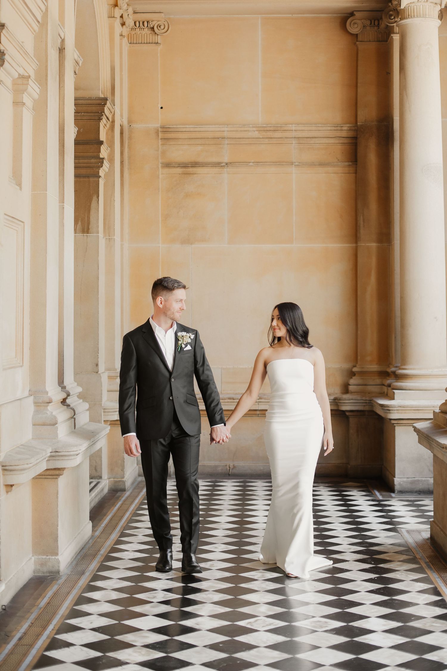 A couple in wedding attire walk hand in hand down an elegant corridor with black and white checkered floors.