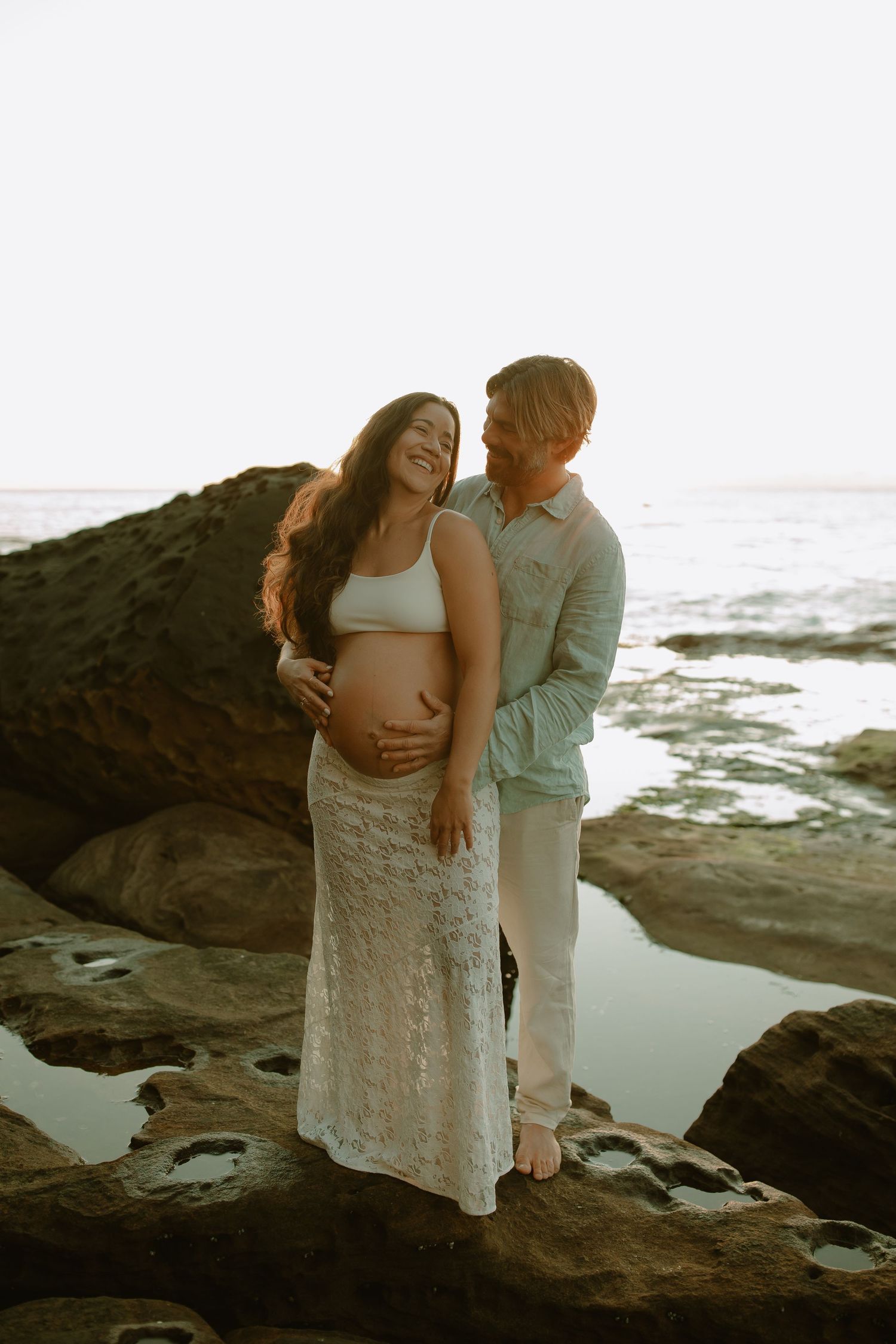 A romantic maternity photo shoot on rocky coastal cliffs at sunset with ocean waves in the background.