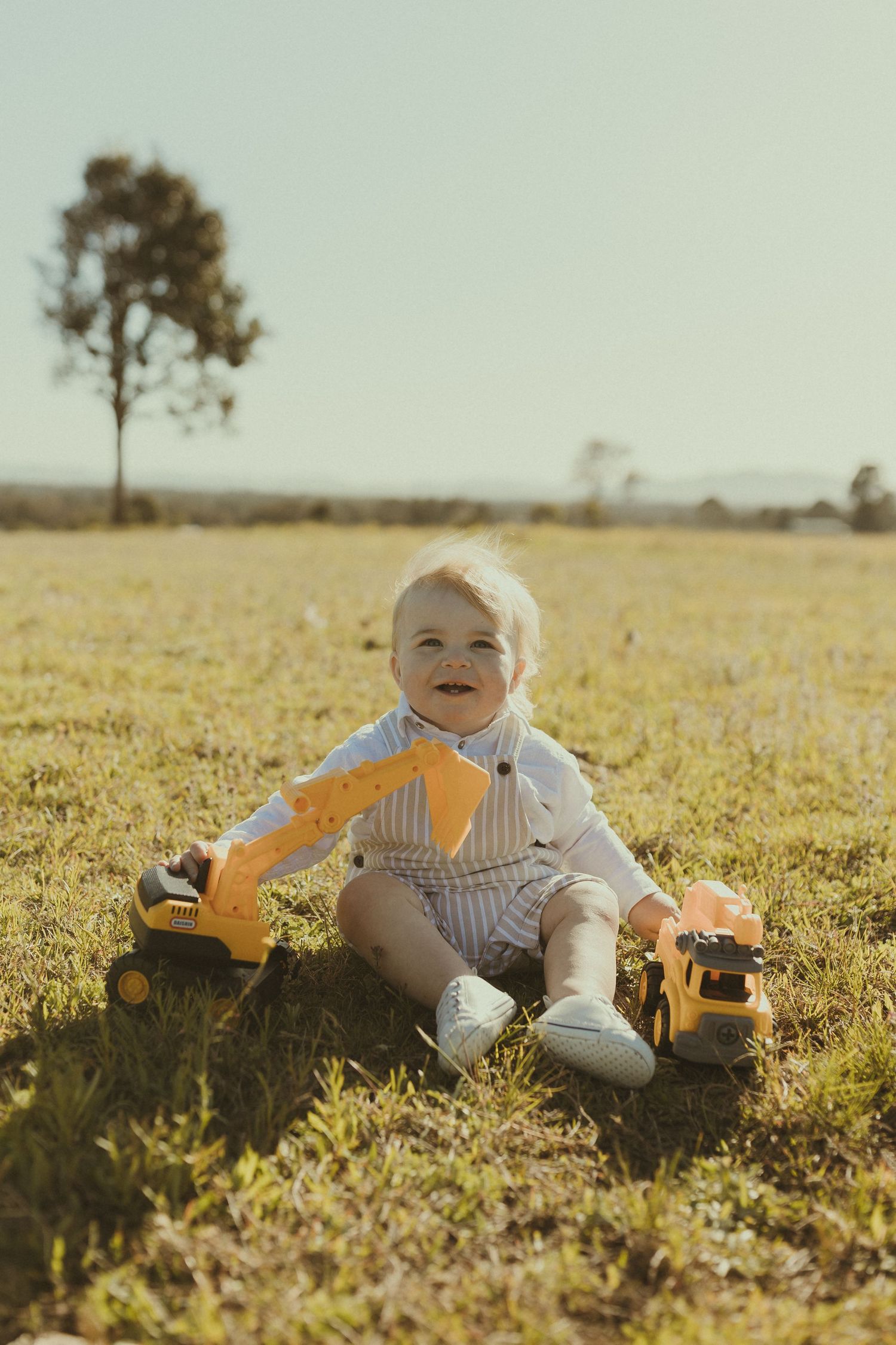 A small child plays while lying in a sunny grass field wearing light colored clothing.
