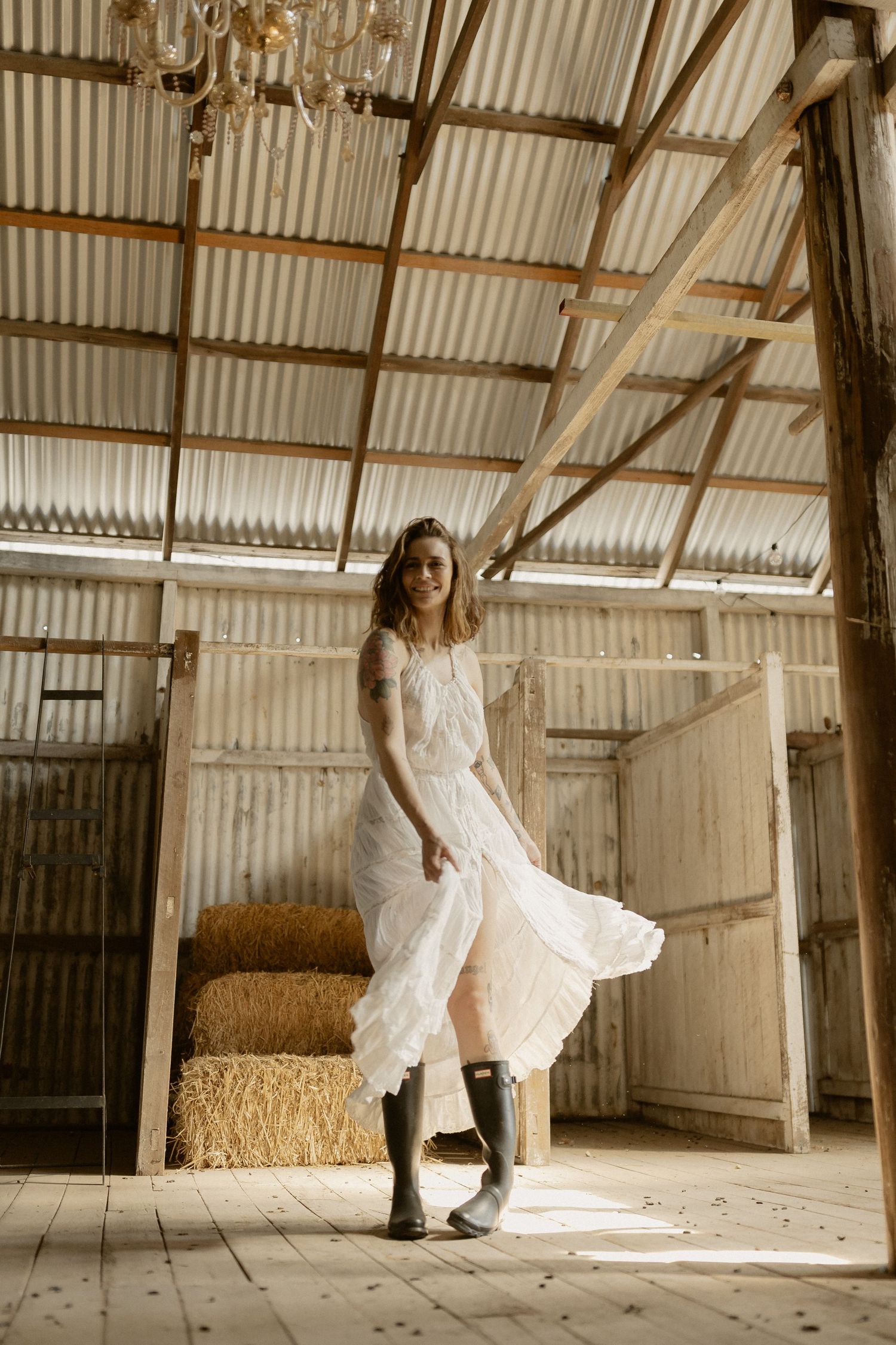 A figure in white twirls elegantly in a rustic barn setting with streaming sunlight and hay bales.