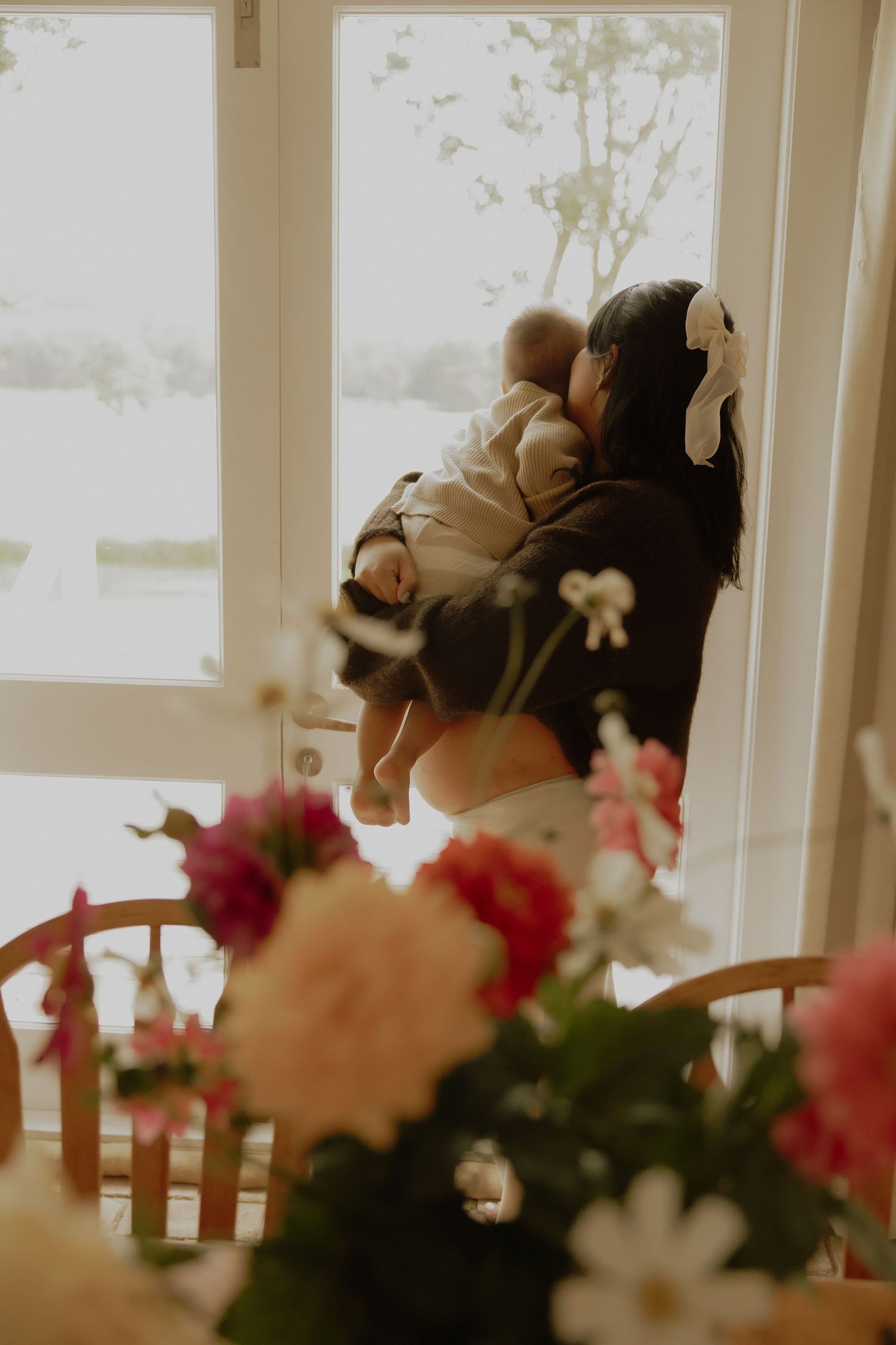 Mother holding newborn baby near bright window with pink flowers in foreground.