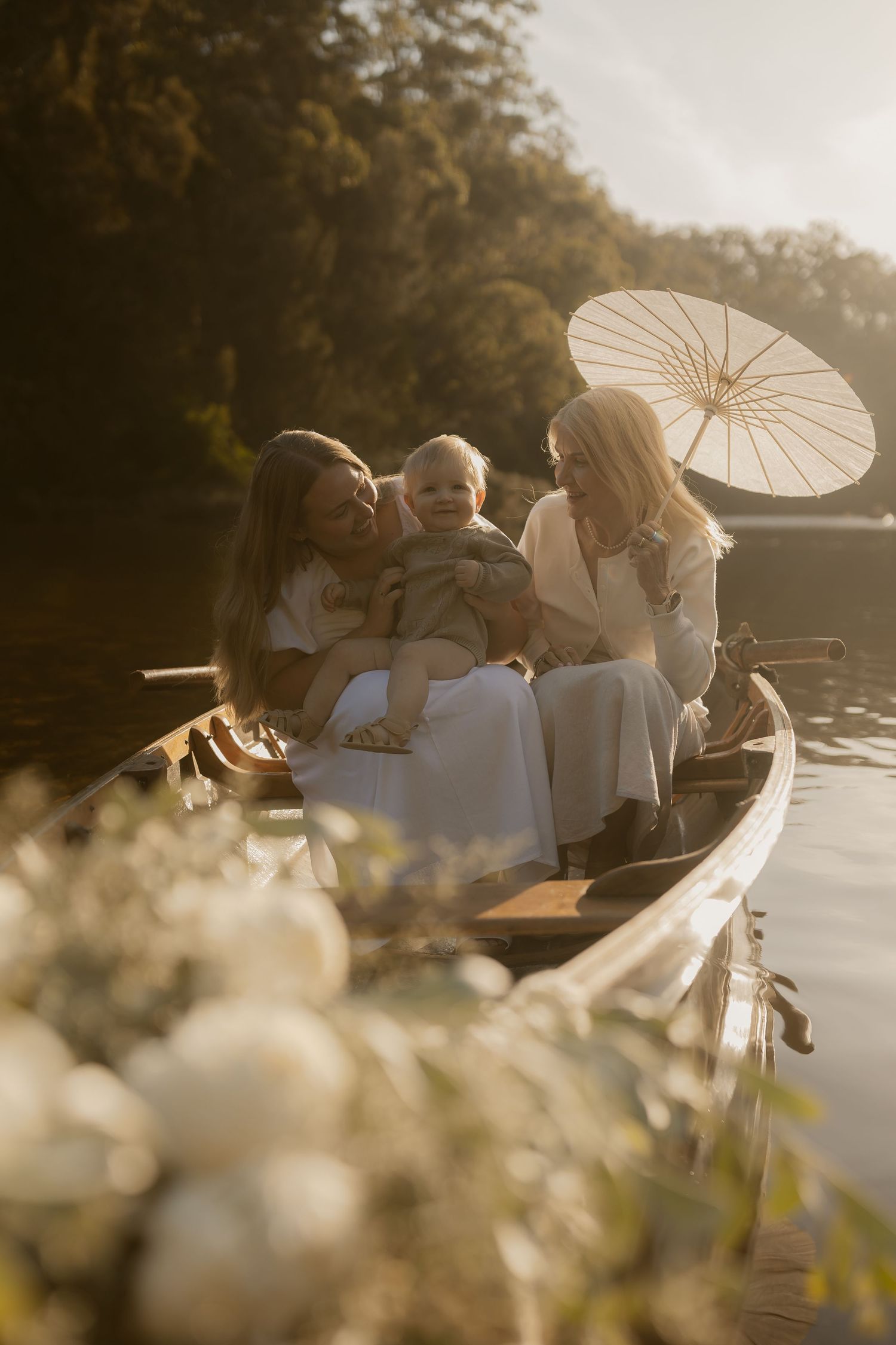 Two women and a baby sit in a wooden boat on a calm lake, surrounded by white flowers and golden sunlight.