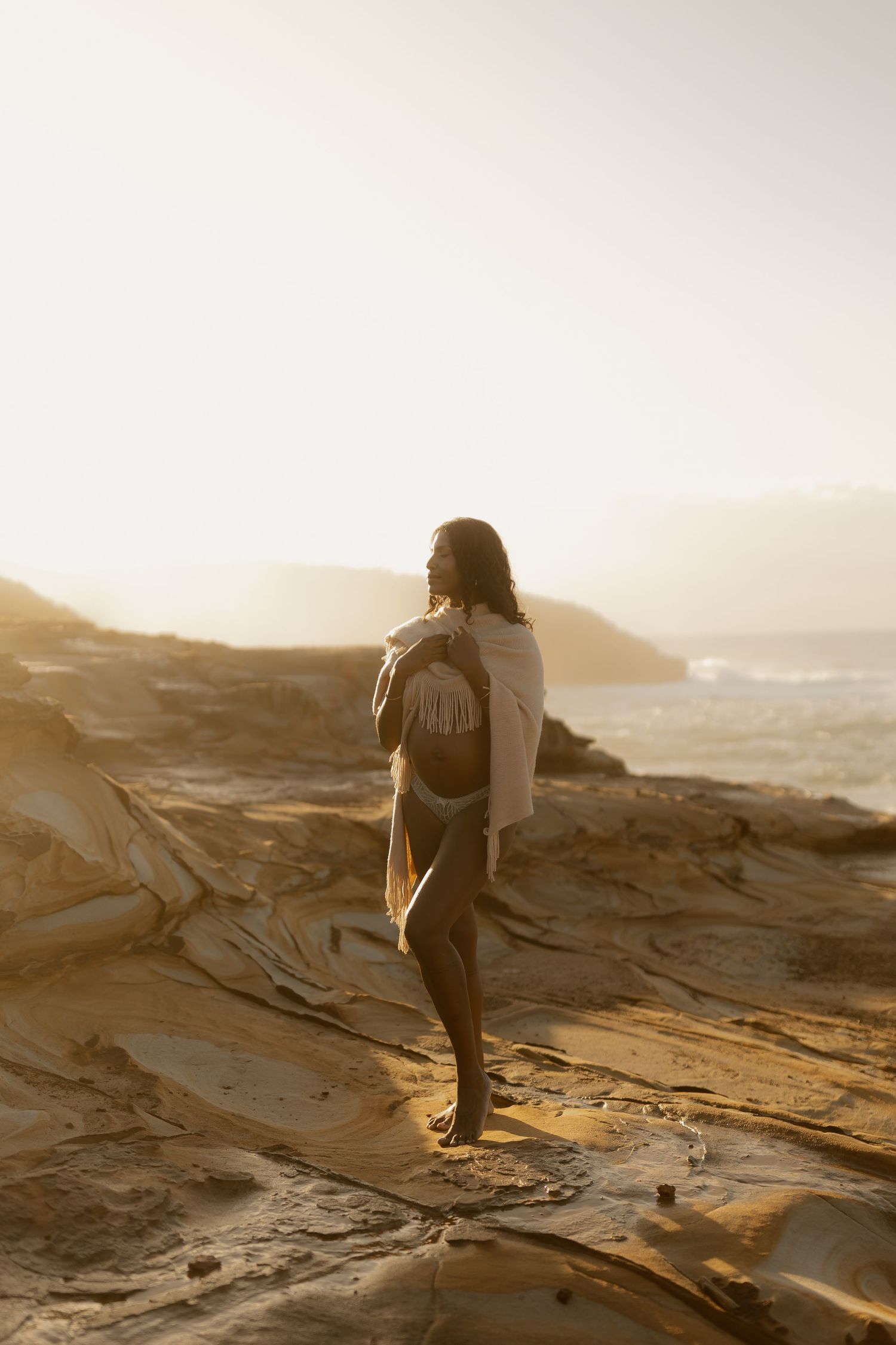 Pregnant woman wrapped in fringed shawl stands on coastal rocks gazing at ocean during sunset.