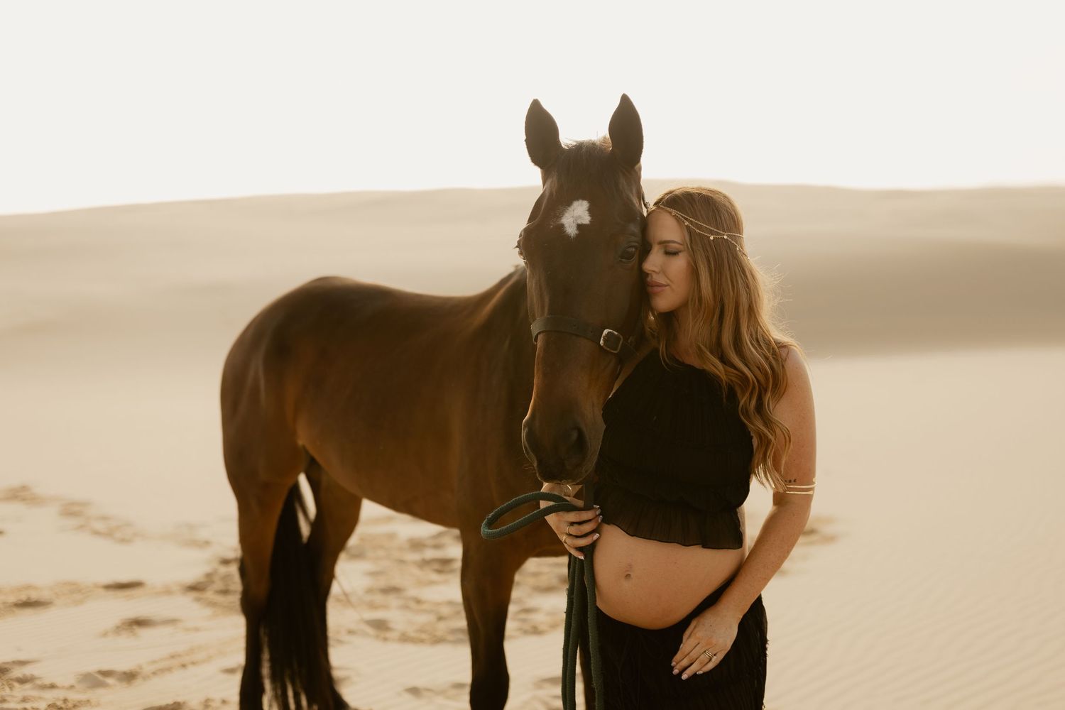 Pregnant woman in black outfit stands beside a brown horse on sandy desert dunes.