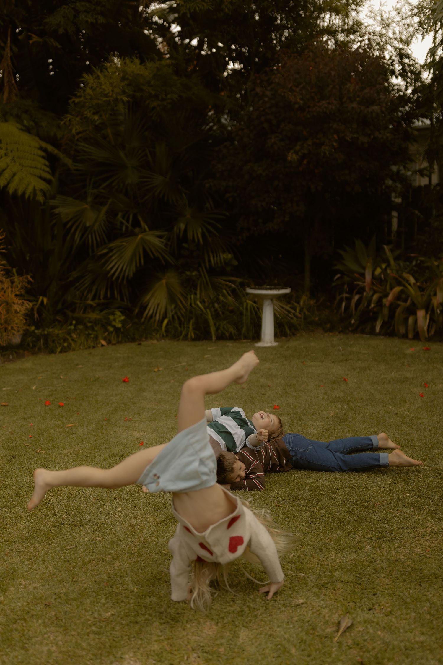 A child does a cartwheel on green grass while another child lies nearby in a lush garden.