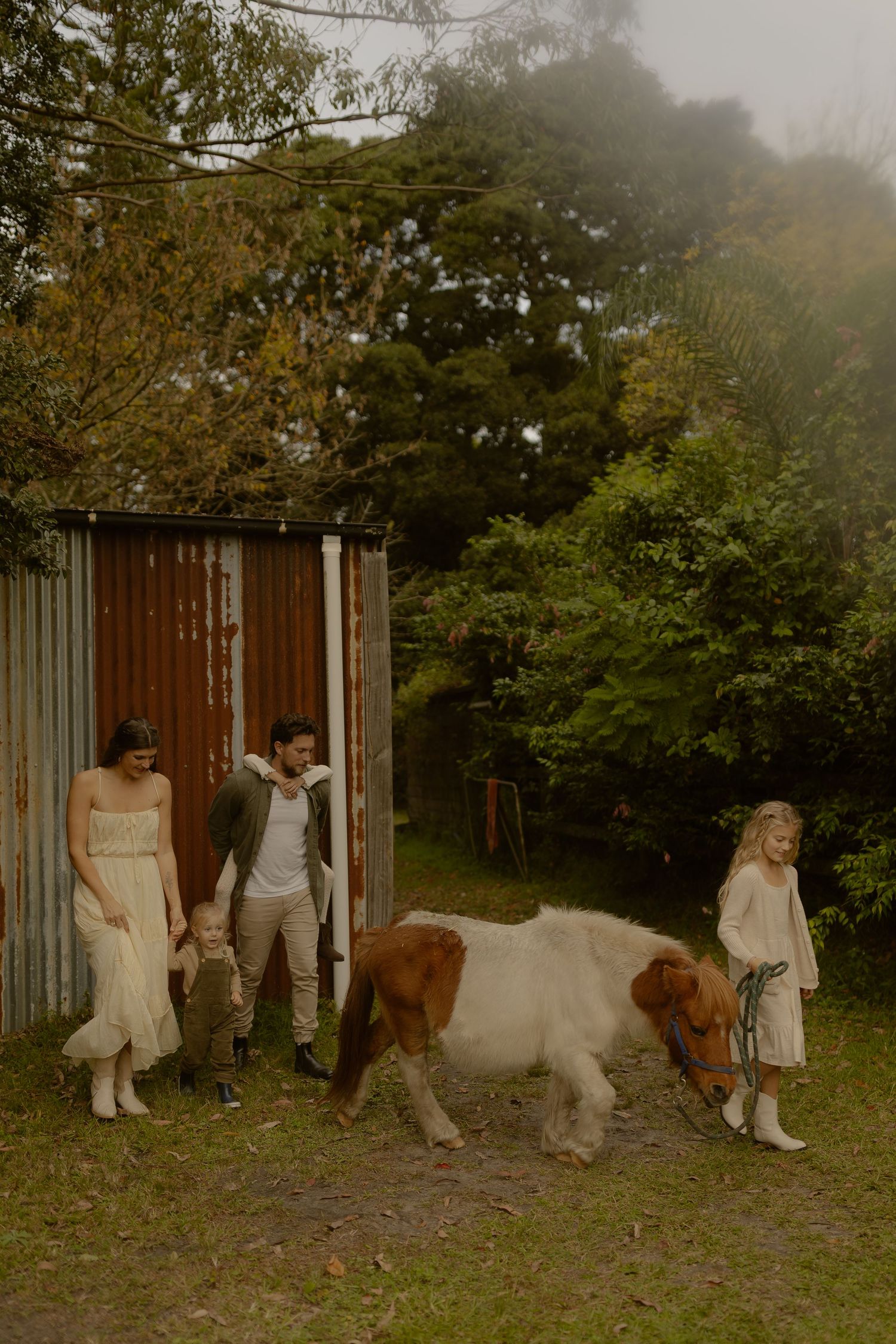 A family with children and a brown and white pony stand near a rustic shed surrounded by greenery.