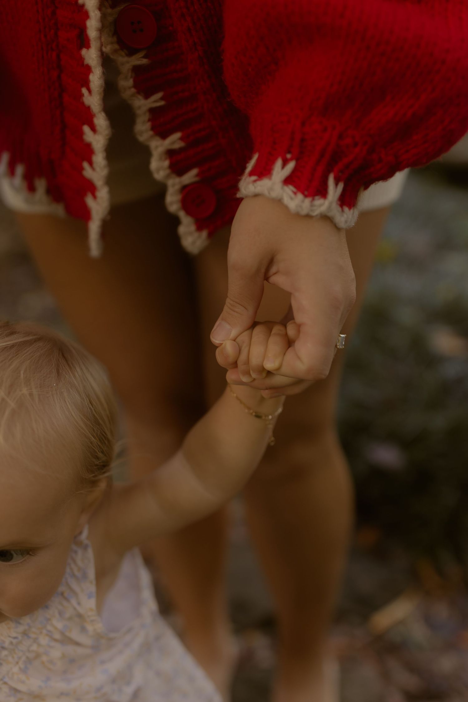 Close-up of mother and baby holding hands, woman wearing red fringed boho dress outdoors.