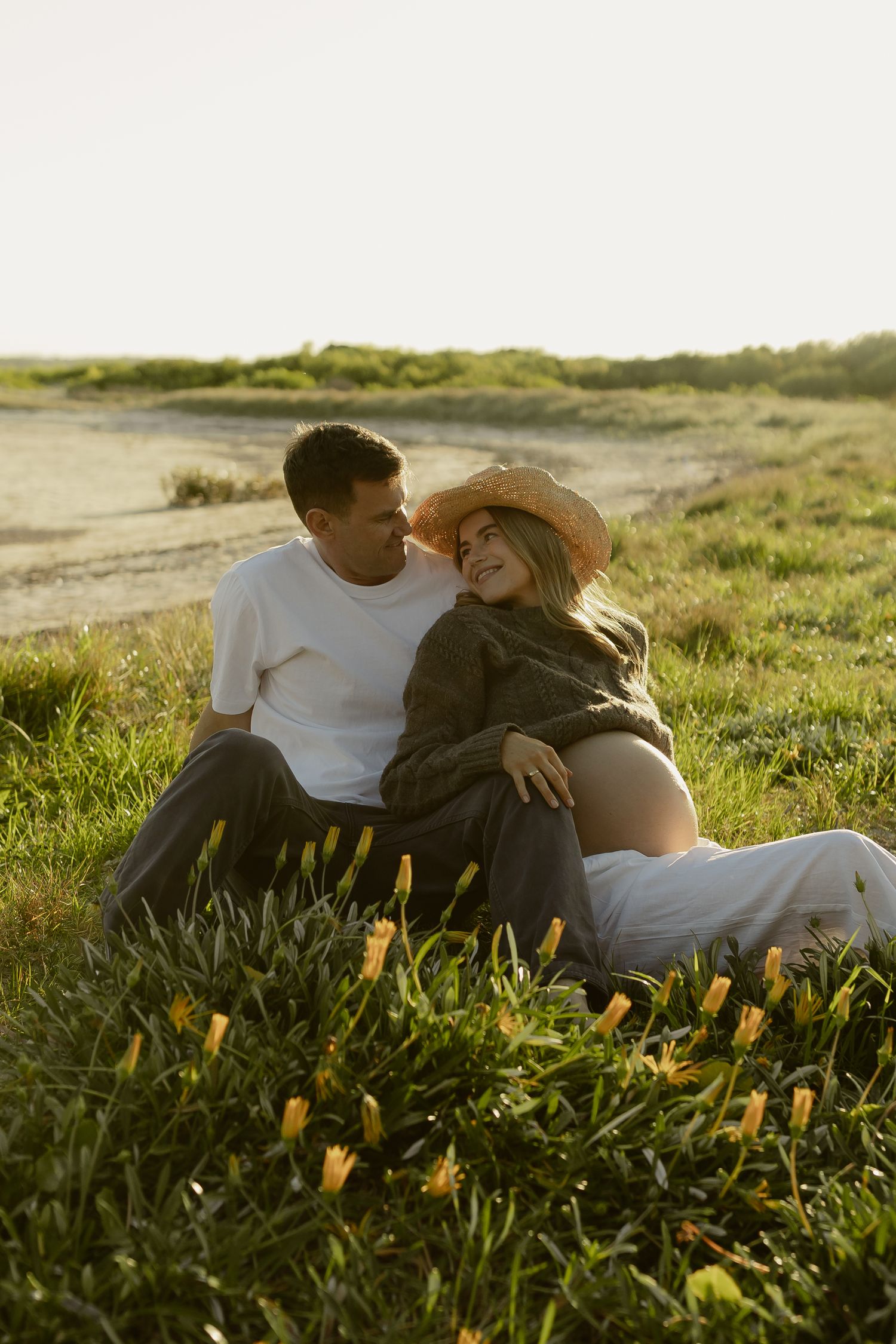 Couple sitting in a sunny green field with yellow flowers, woman wearing straw hat cradling pregnant belly.