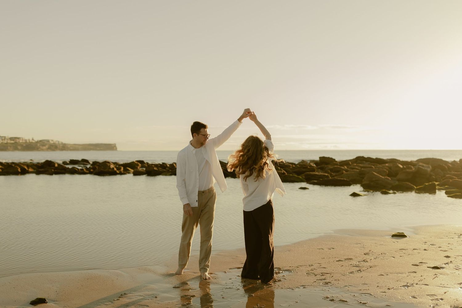Couple dances together on wet sandy beach at sunset during romantic maternity photo session.