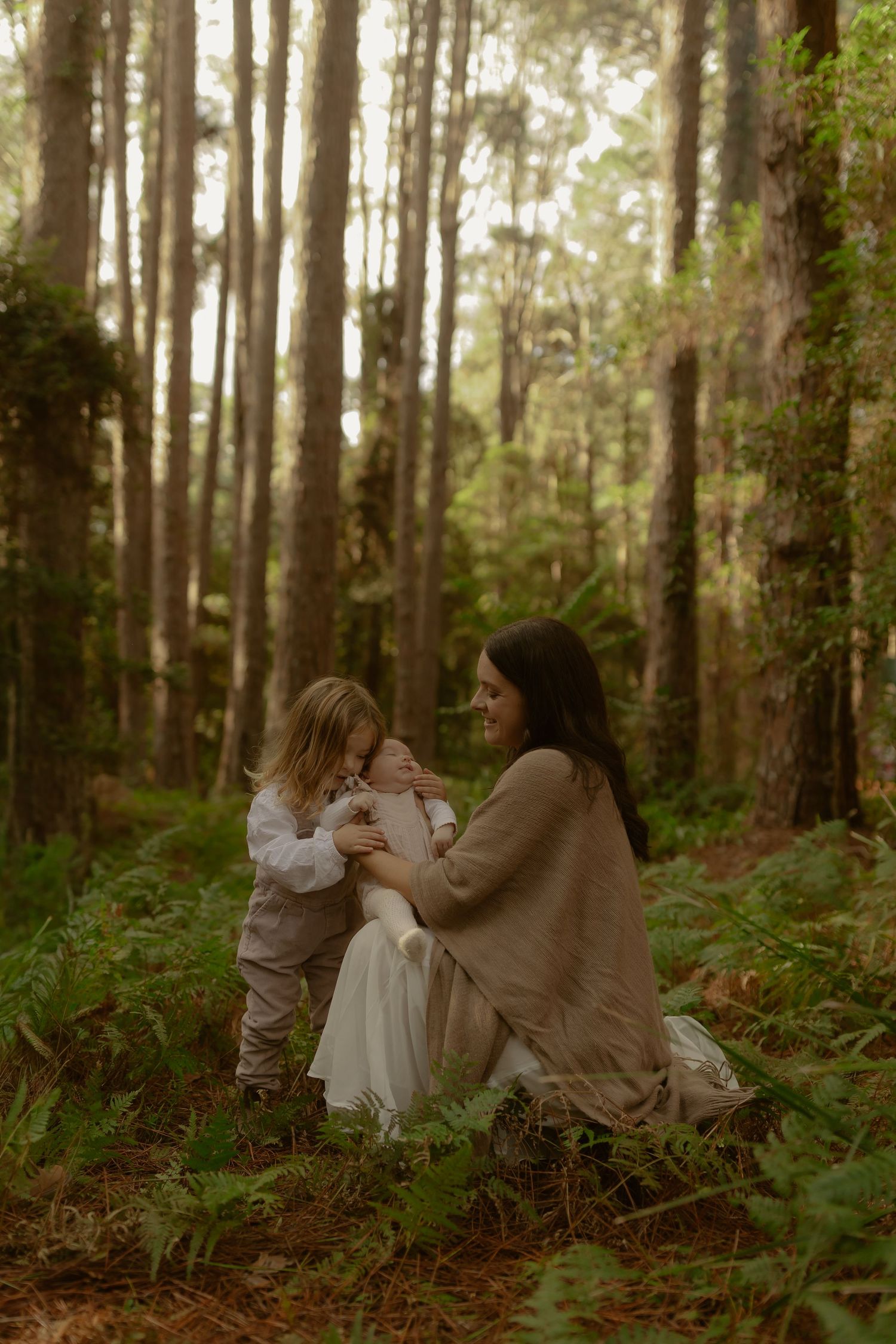 Mother sits in forest holding newborn while young child leans in lovingly.